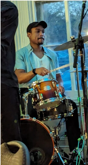 Young man playing drums in a room with large windows and blue sky visible outside.