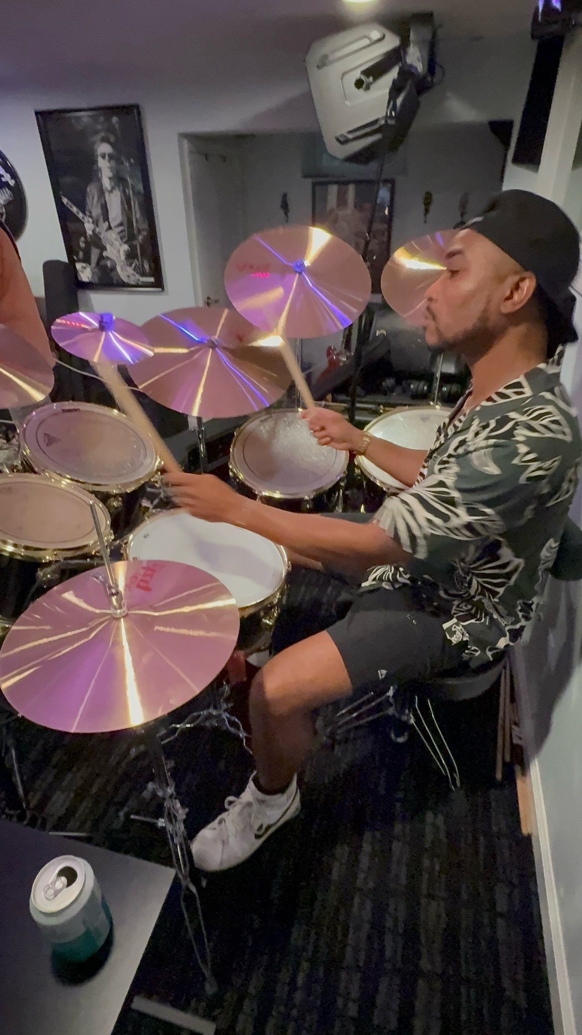 A man wearing a black cap and patterned shirt playing a drum set in a room decorated with posters and music equipment.