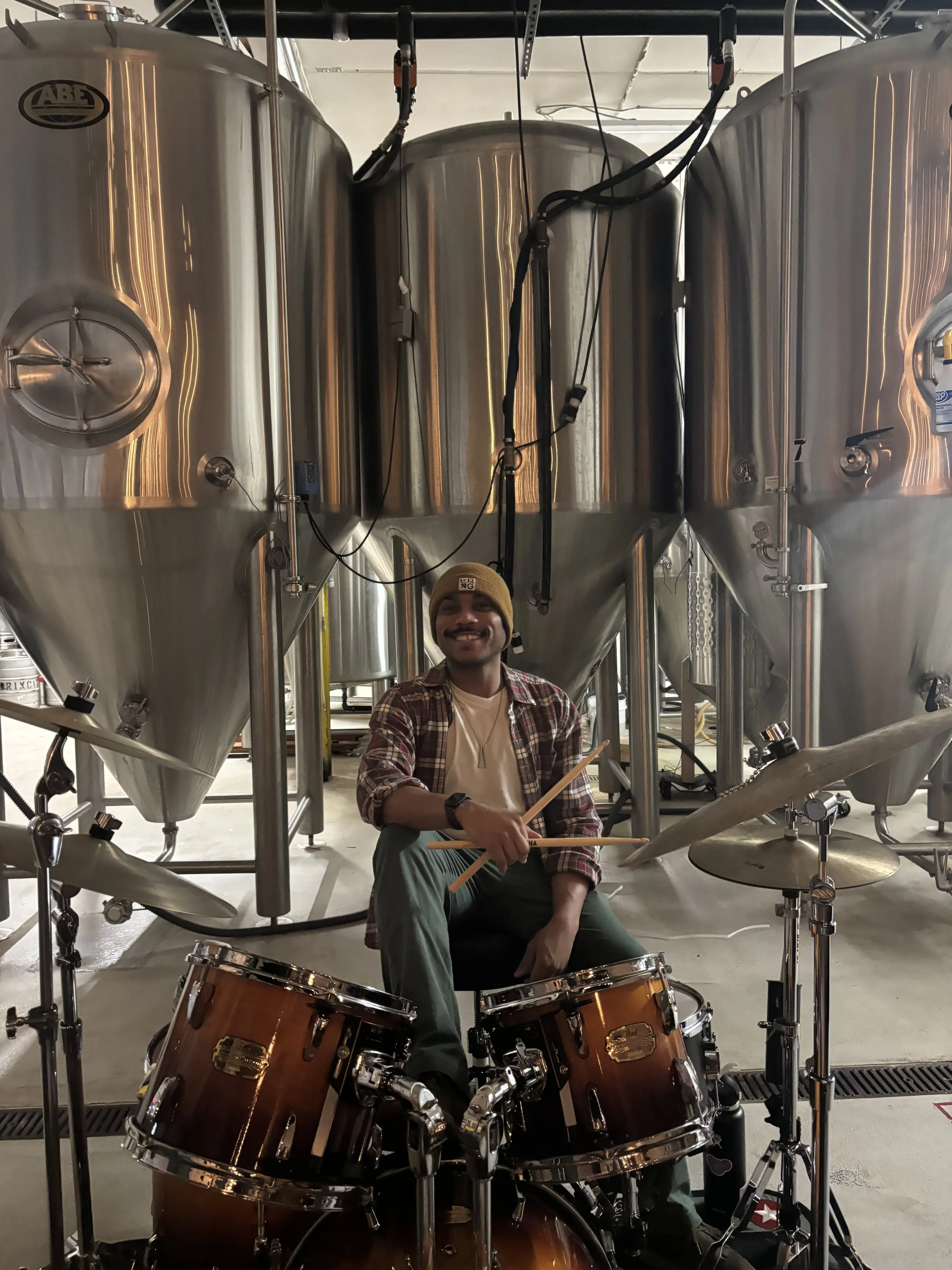 A person smiling and sitting behind a drum set holding drumsticks, in a brewery with large stainless steel fermentation tanks in the background.
