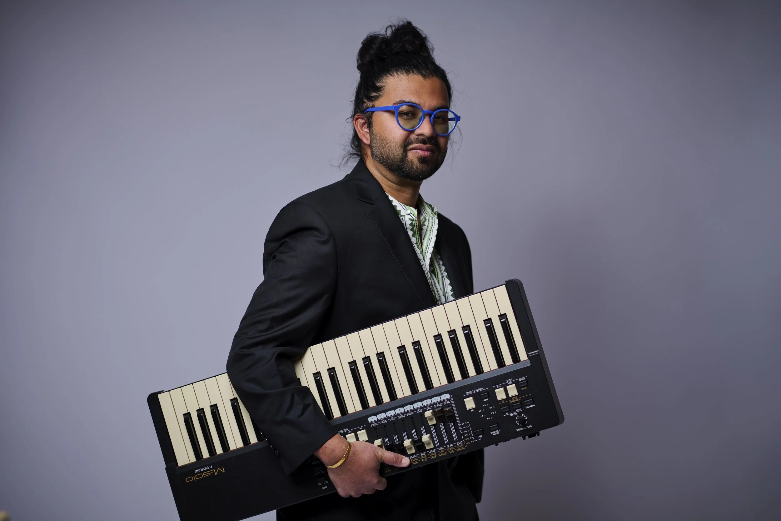 A man with long curly hair, glasses, and a beard holding a vintage synthesizer keyboard in front of a gray background.