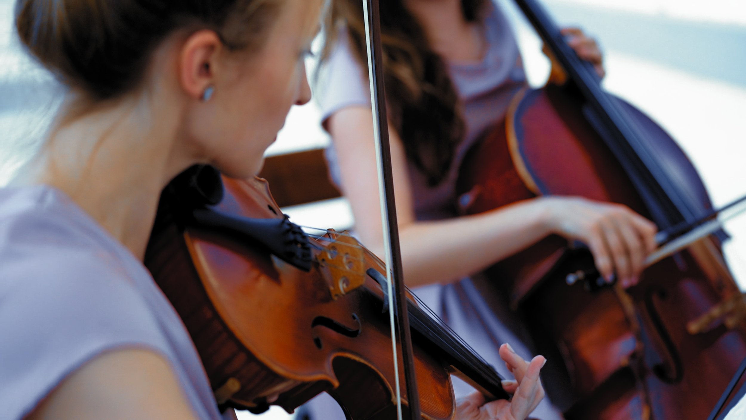 Two women are playing violins outdoors, focused on their instrument, with a mirror reflecting their side view.