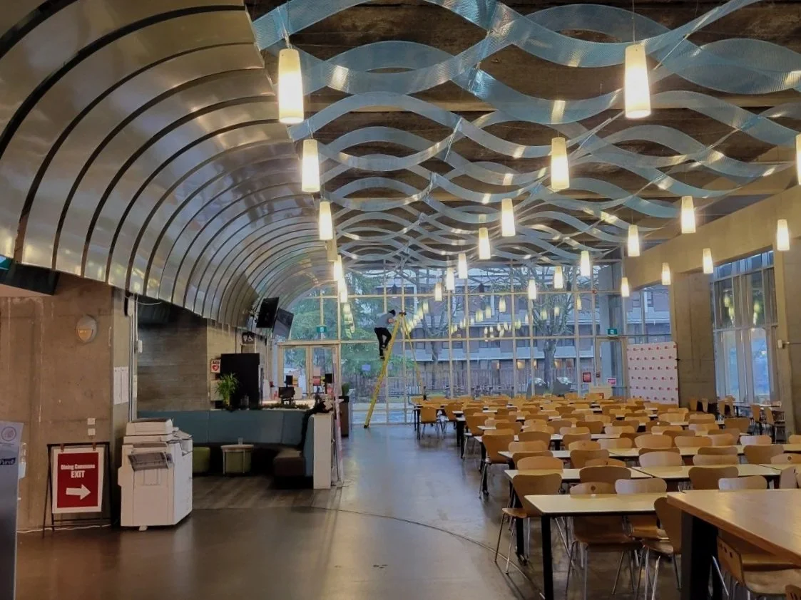 Indoor space with ceiling decorated with wavy patterns and hanging lights, several tables and chairs arranged in rows, a person on a ladder working near the windows, and various electronic equipment near a reception area.
