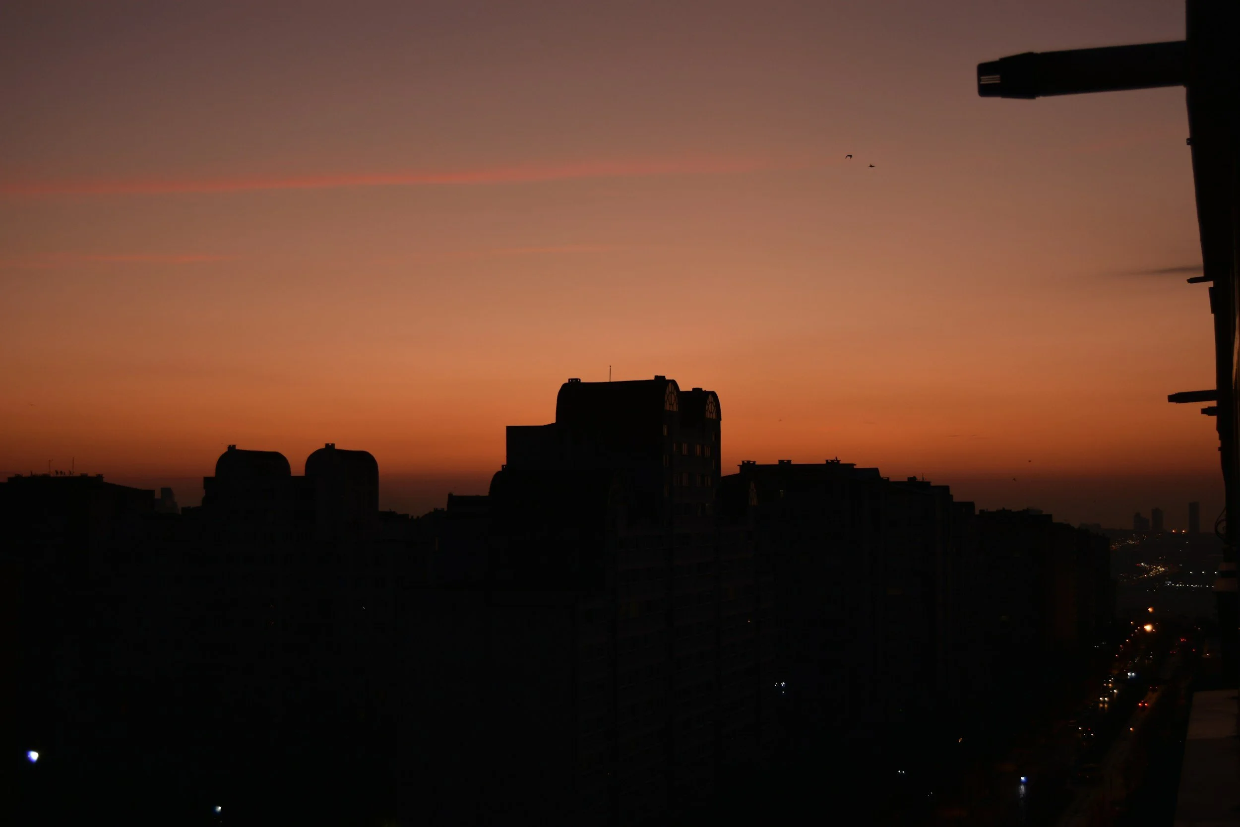 Silhouettes of tall buildings against a sunset sky with a gradient of orange and purple hues, and a few distant birds in the sky.