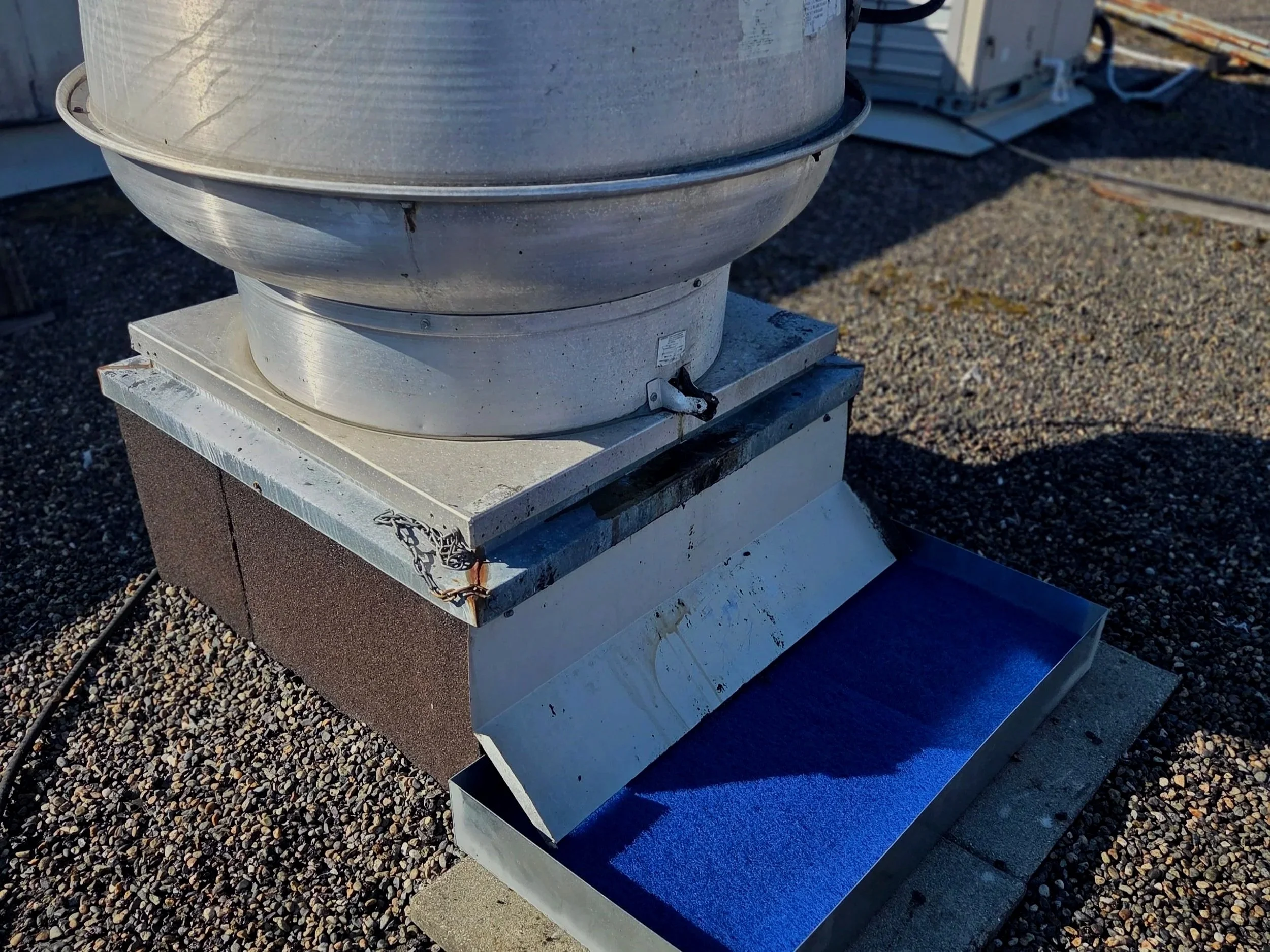 Close-up of a rooftop HVAC exhaust vent with a metal protective shelter, installed on a gravel roof surface.