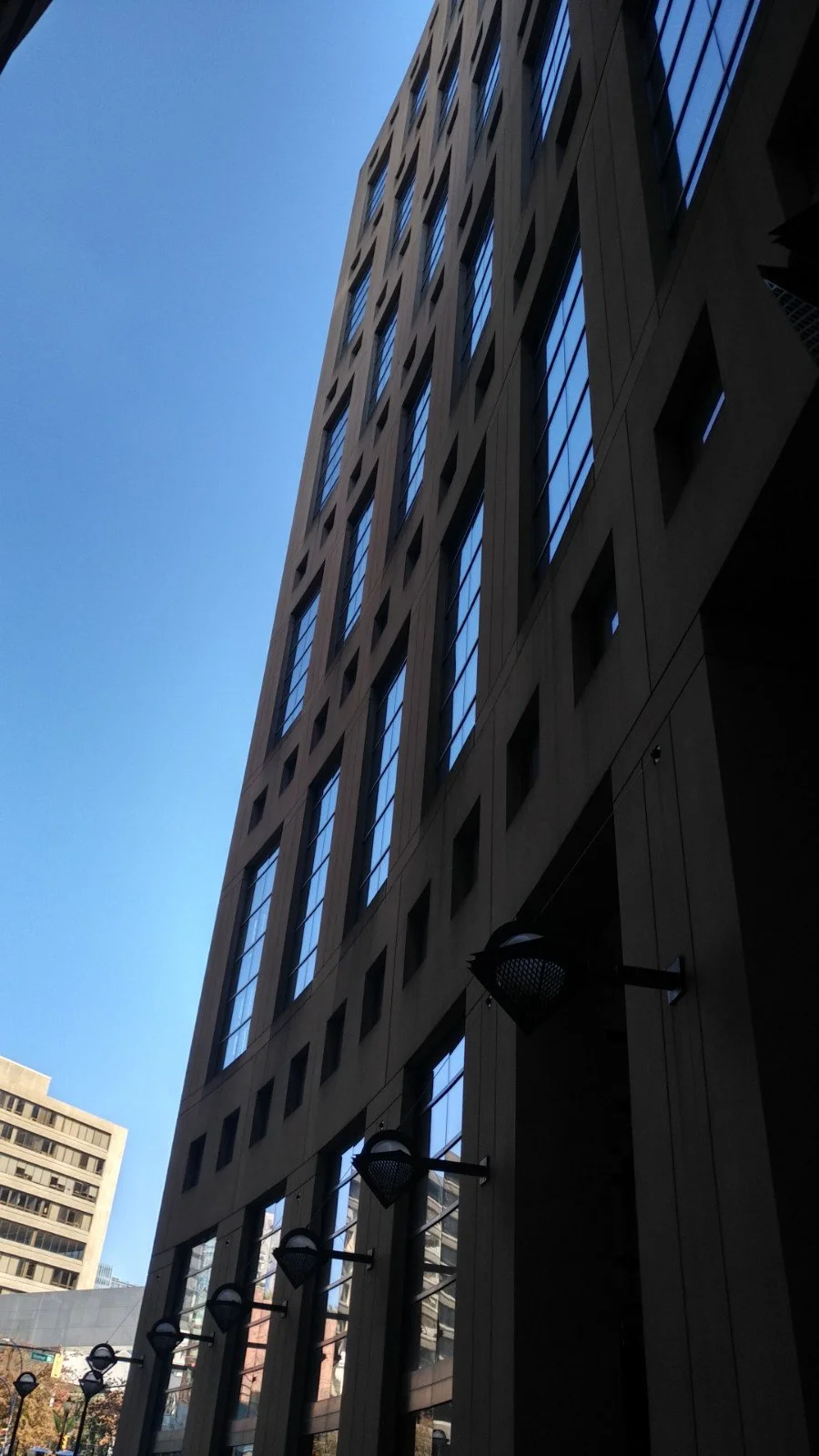 Looking up at a tall modern building with rectangular windows and a beige facade, blue sky in the background, and street lamps along the sidewalk.