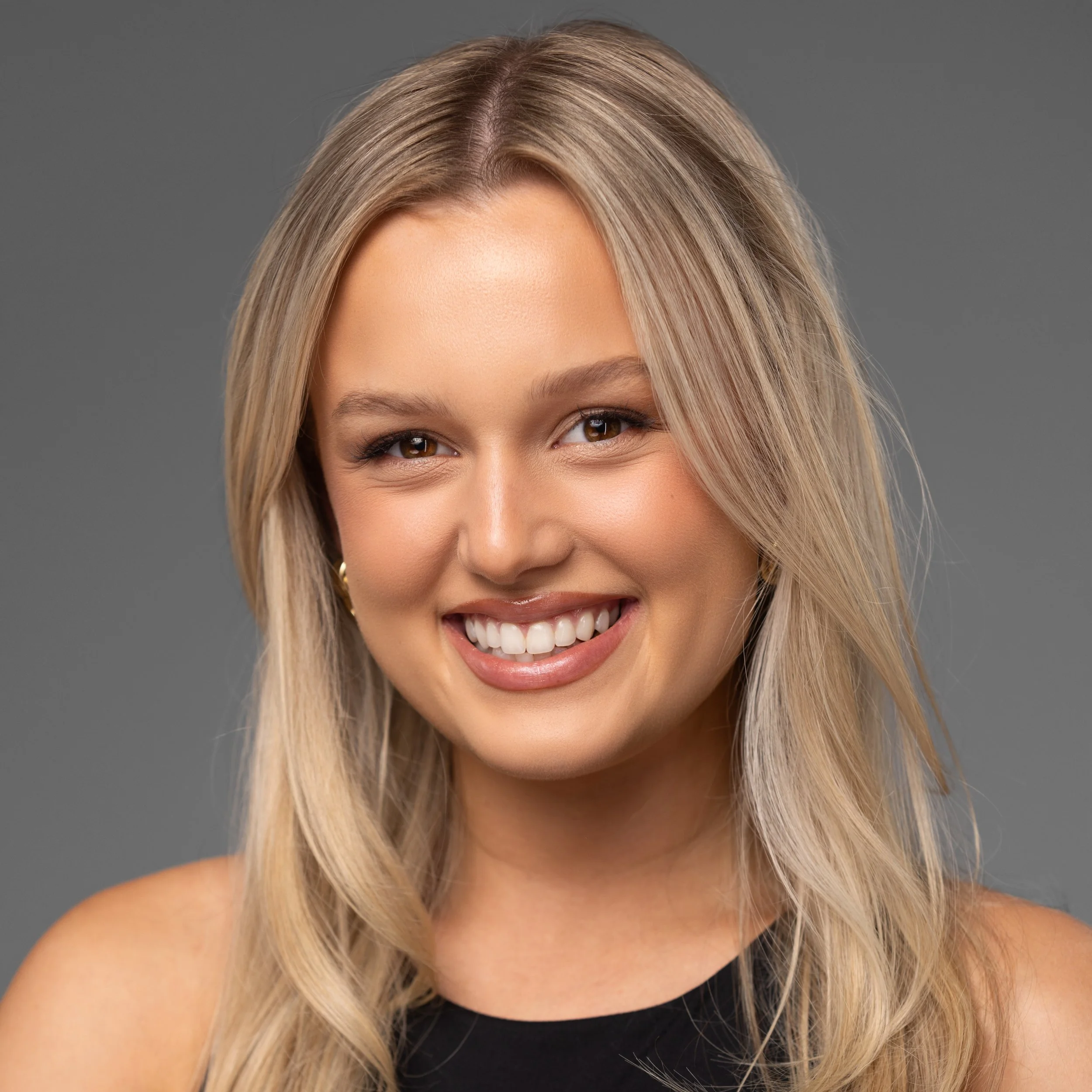 A smiling woman with blonde hair, wearing makeup, earrings, and a black top, against a gray background.