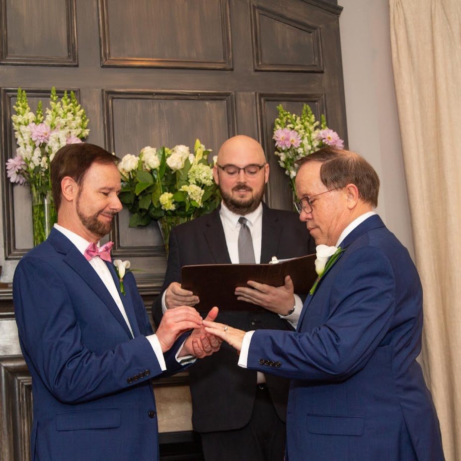 Two men in blue suits exchanging wedding rings during a wedding ceremony, officiant holding a book behind them, with a floral arrangement of pink, white, and green flowers on the dark wood-paneled wall.