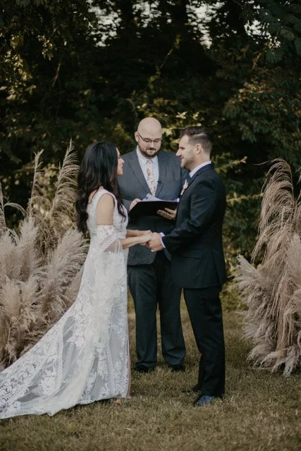 A couple getting married outdoors, holding hands and facing each other, with a officiant standing behind them, in front of tall grass and trees.