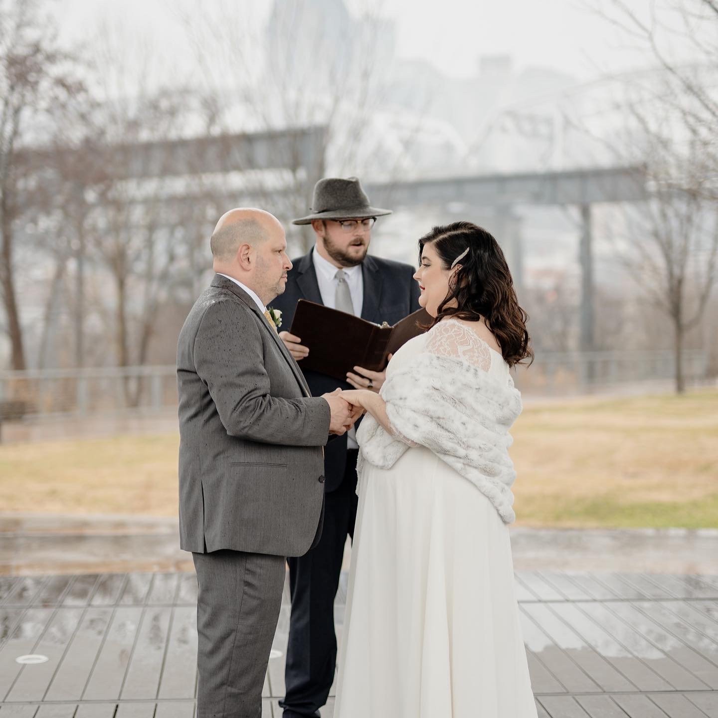 A couple getting married outdoors with an officiant. The groom is in a gray suit, and the bride is in a white dress with a fur shawl. They are holding hands and looking into each other's eyes, with an officiant reading from a book behind them.