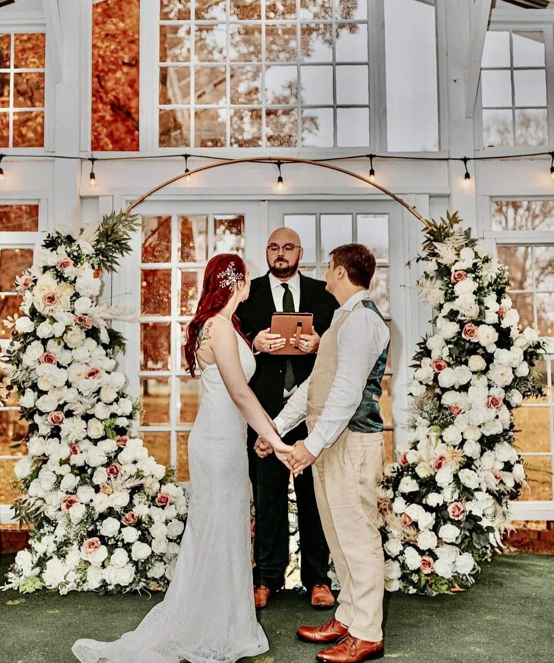 A wedding ceremony with a bride and groom holding hands, standing before an officiant, inside a glass-enclosed venue decorated with large white and pink floral arrangements and string lights.