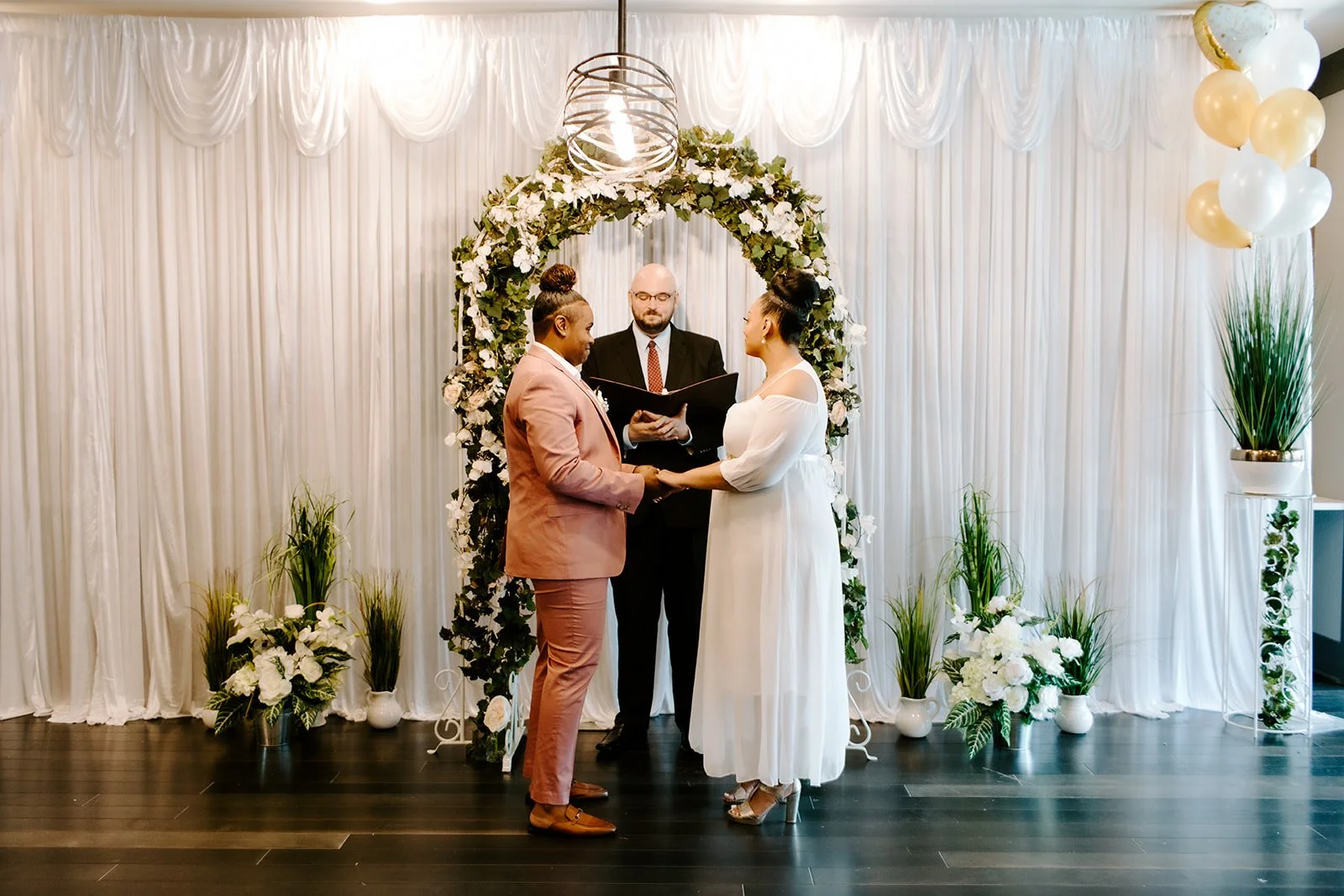 Couple getting married, holding hands, standing before an officiant in front of a decorative floral wedding arch, with white drapes and potted plants in the background.