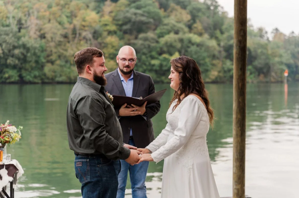 A couple getting married outdoors by a lake, holding hands and smiling at each other, with an officiant standing behind them.