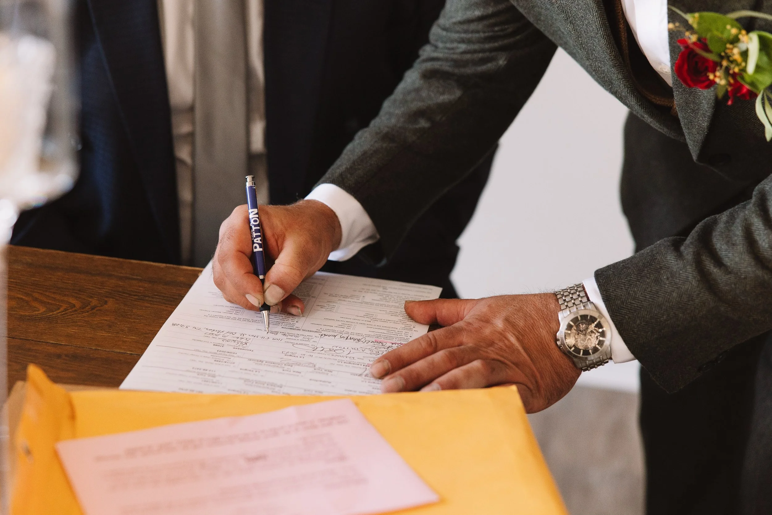 Person in a suit signing a document on a wooden table with a yellow folder in the foreground.