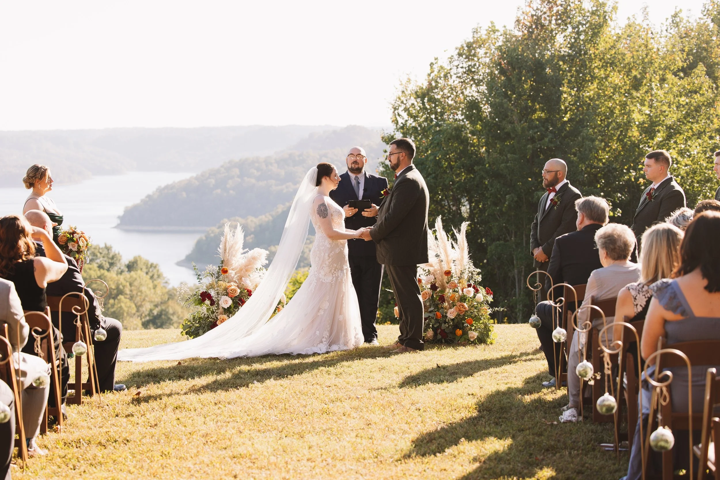 Outdoor wedding ceremony with the officiant, bride, and groom standing before an audience, overlooking a river and forested hills, during daytime.