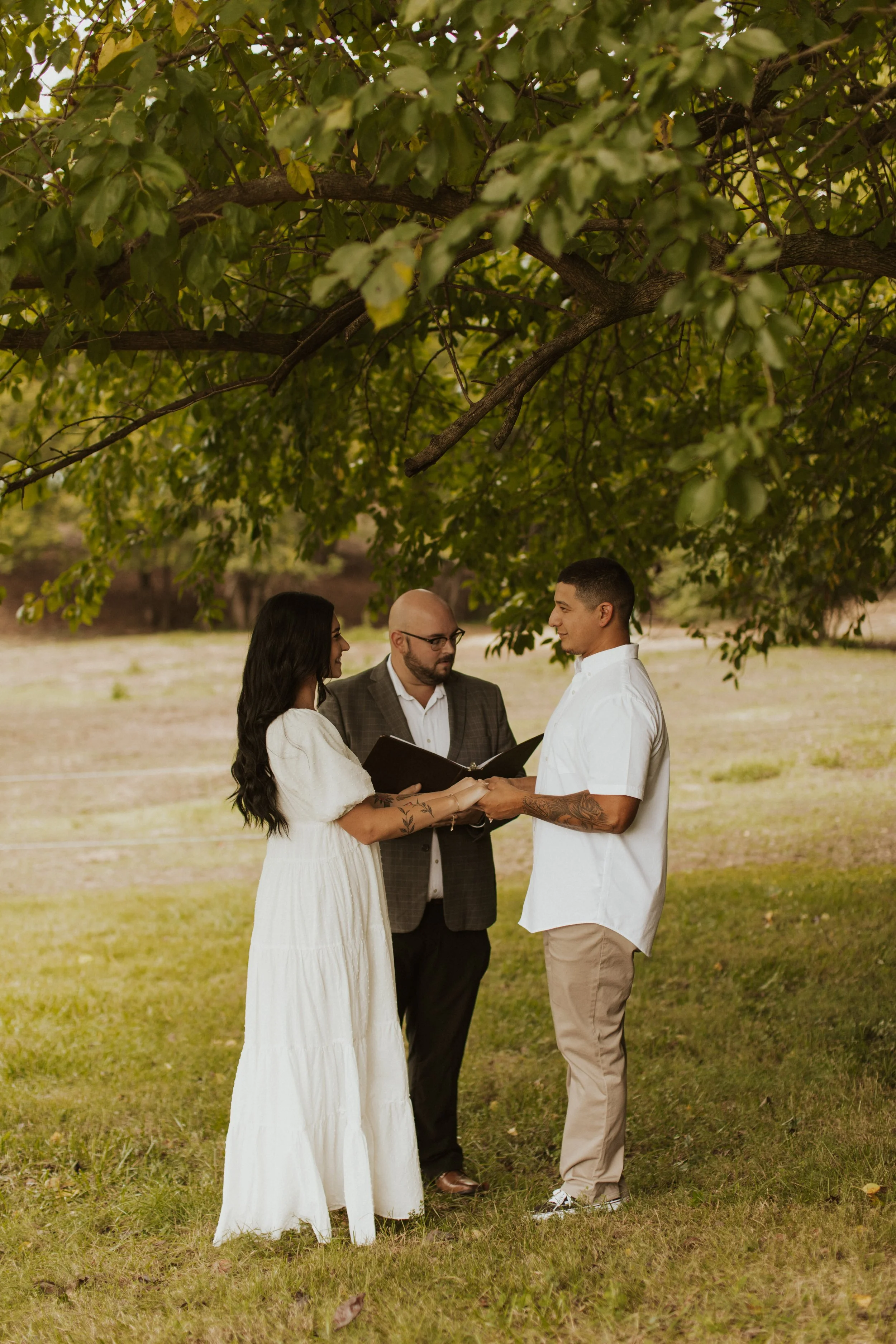 A couple is getting married outdoors under a large tree, with a officiant reading vows. The bride is wearing a white dress, and the groom is in a white shirt and beige pants. They are facing each other holding hands.