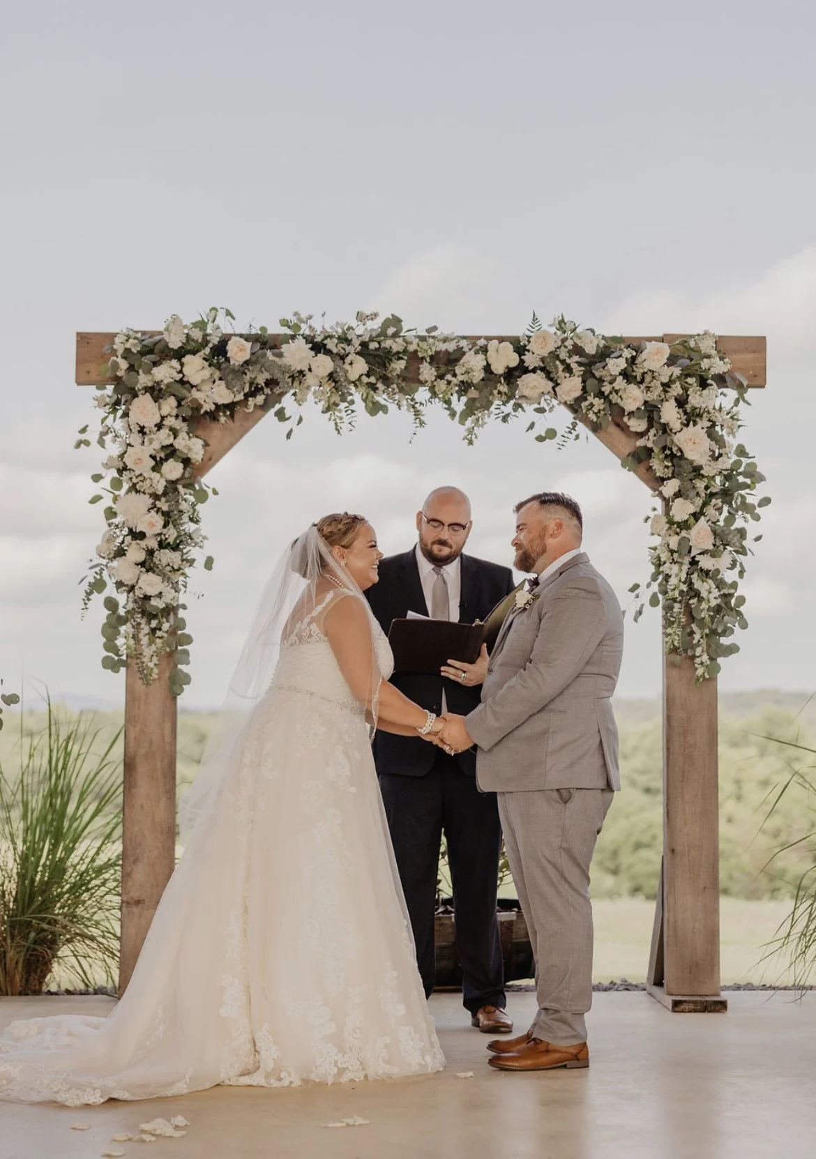 A couple getting married, holding hands and facing each other, standing under a wooden arch decorated with flowers. An officiant is standing behind them, reading from a book. The setting appears to be outdoors with a cloudy sky and green landscape in the background.