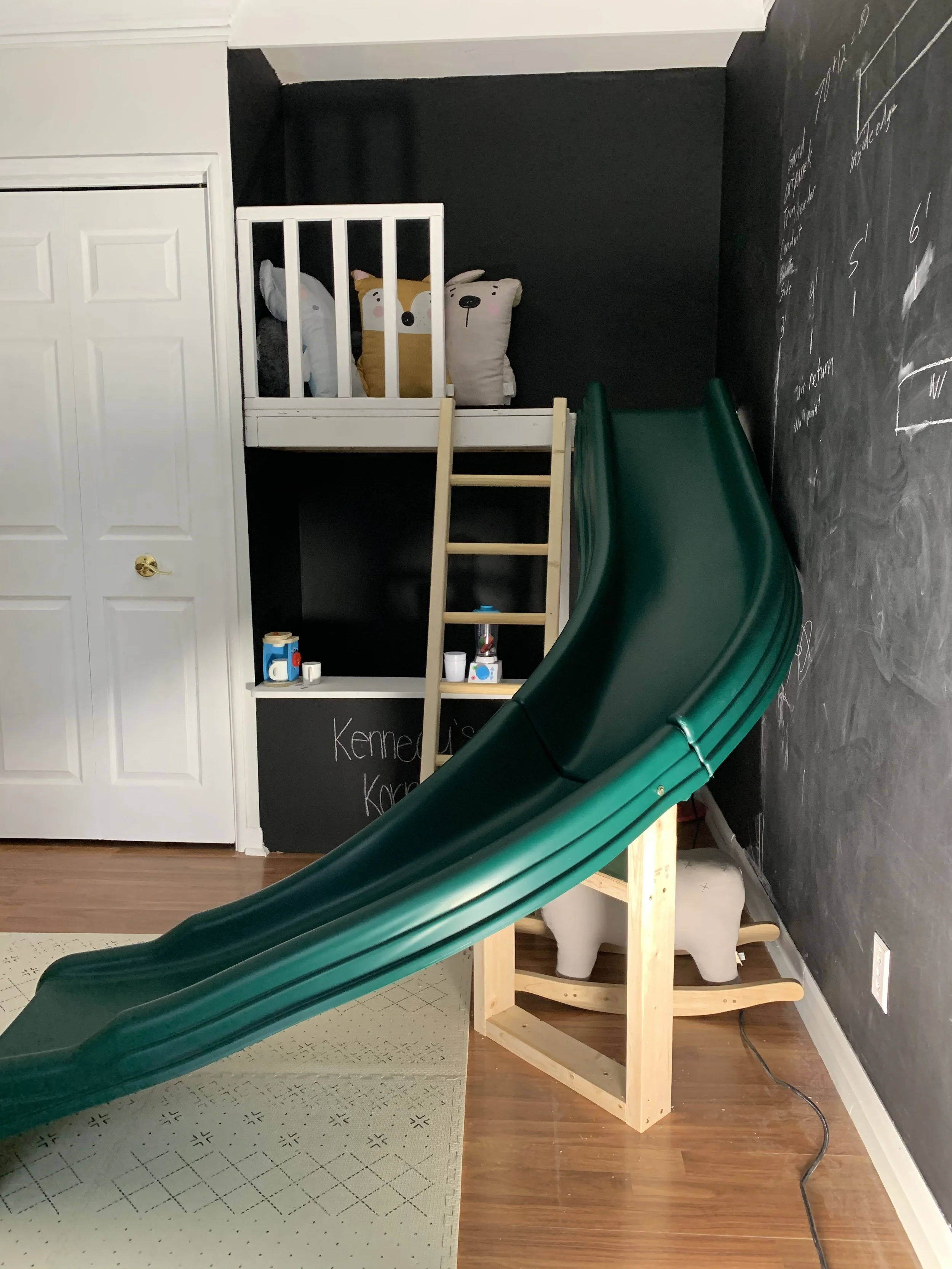 Indoor play area with a green slide attached to a wooden structure, a loft bed with stuffed animal pillows, black chalkboard wall with writing, and white door.