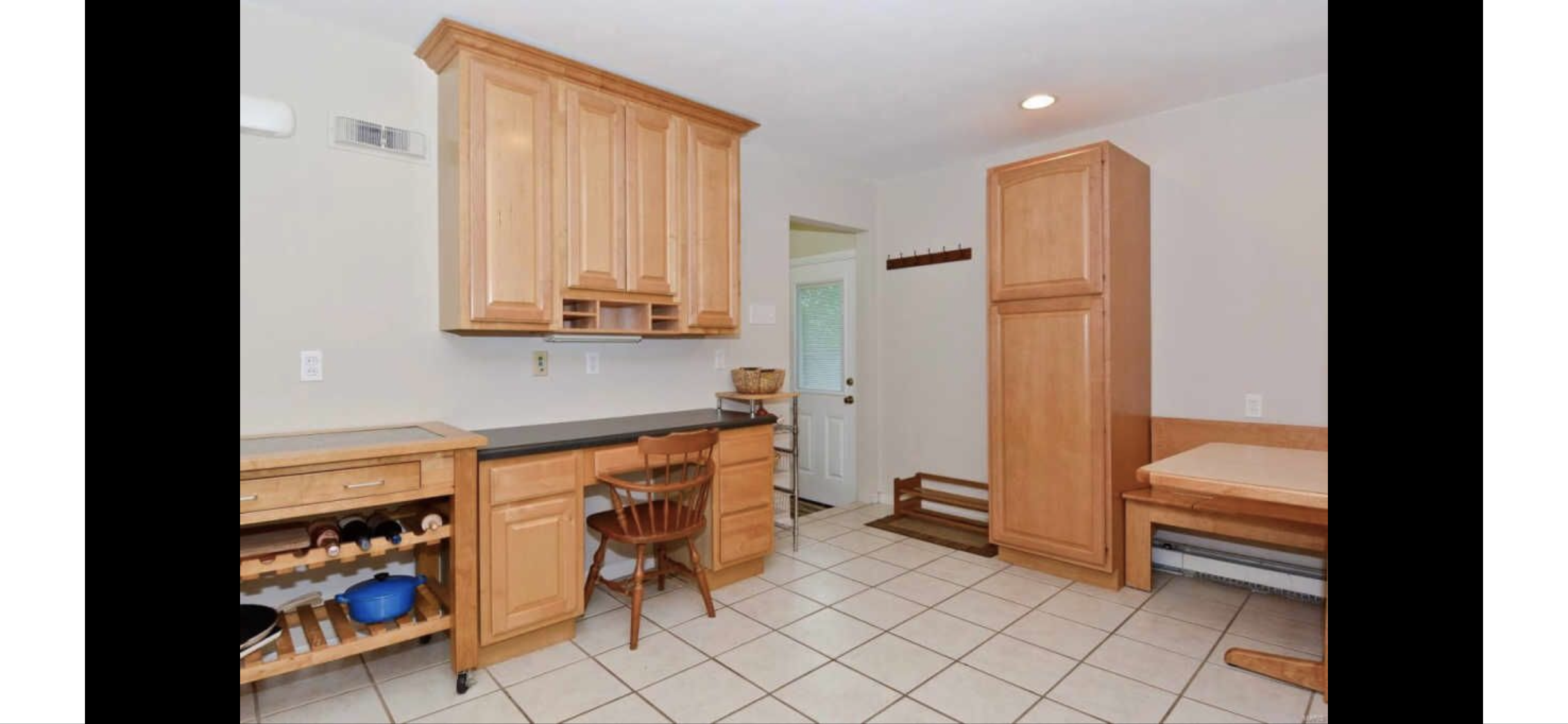 Kitchen with light wood cabinets, tile floor, small desk area with chair, wine rack, small table, and bench with storage.