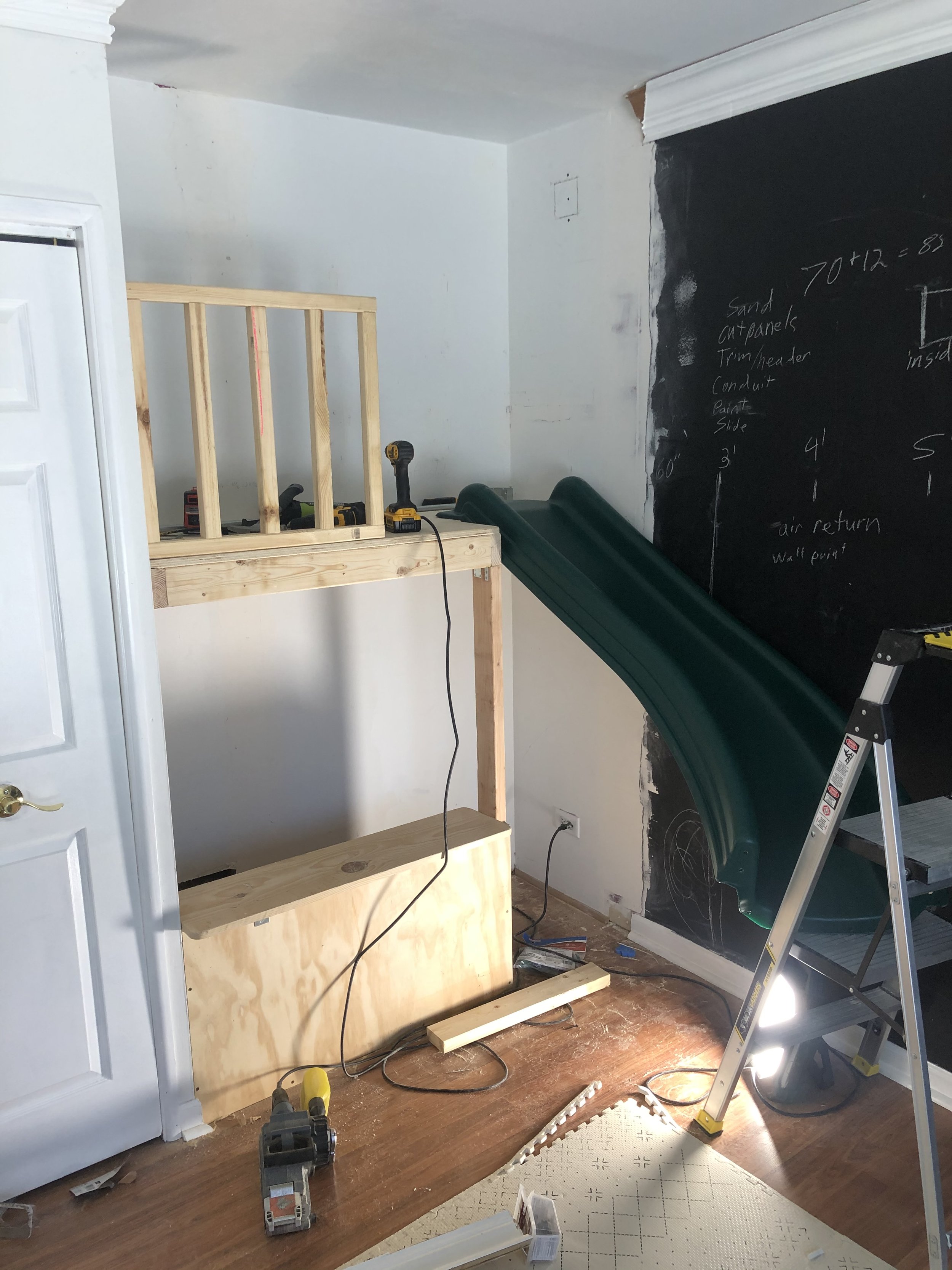 A room under construction with a wooden framework for a small loft or play area, a green slide attached to the loft, construction tools like a drill and power saw, a black chalkboard wall with writing and drawings, and a step ladder.