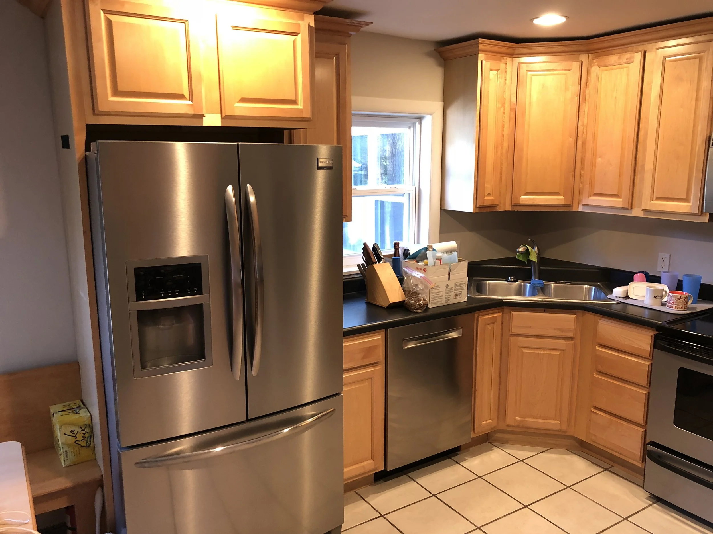 A kitchen with wooden cabinets, a stainless steel refrigerator, a double sink under a window, and various kitchen items on the black countertop.