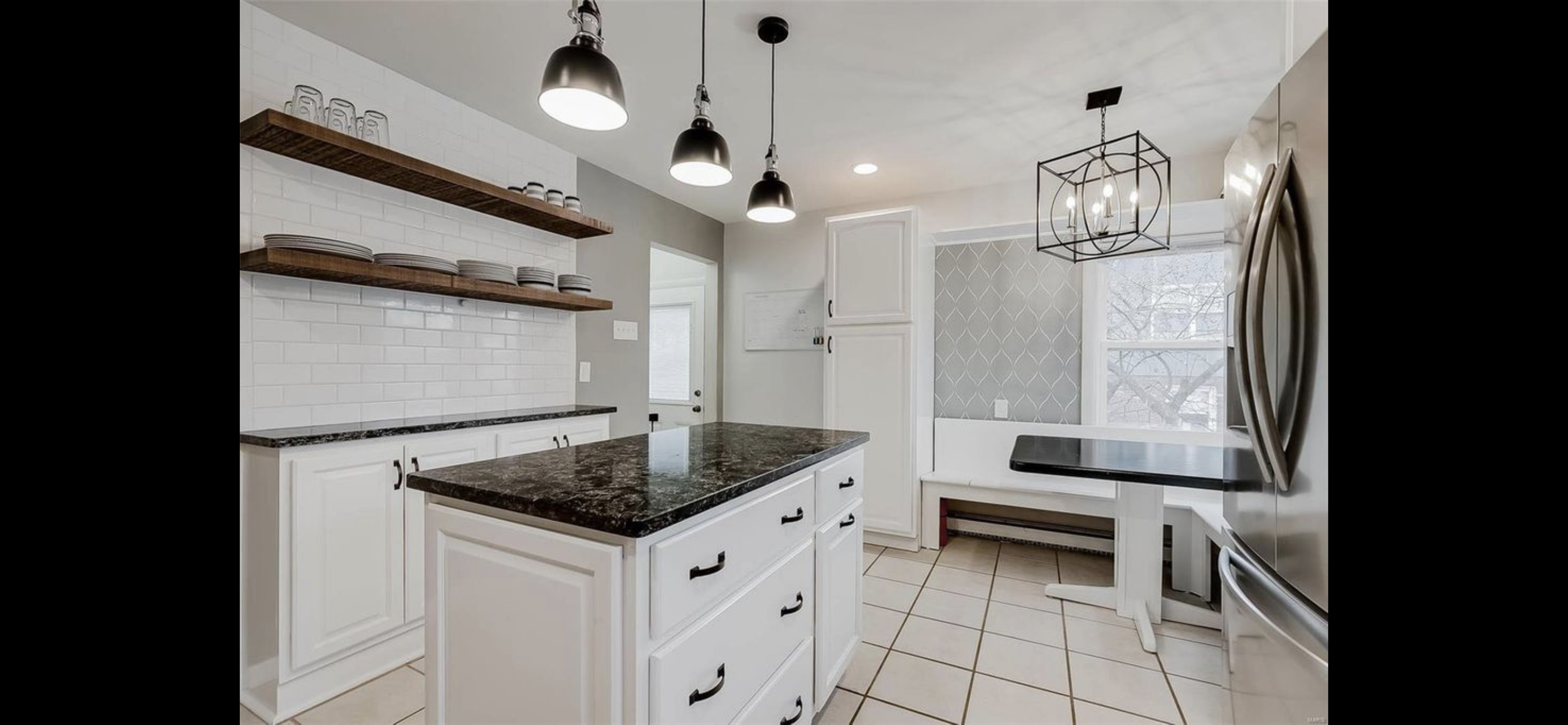 Modern kitchen with white cabinets, black granite countertops, open wooden shelves with dishes, stainless steel refrigerator, black pendant lights, and a large window with patterned wallpaper.