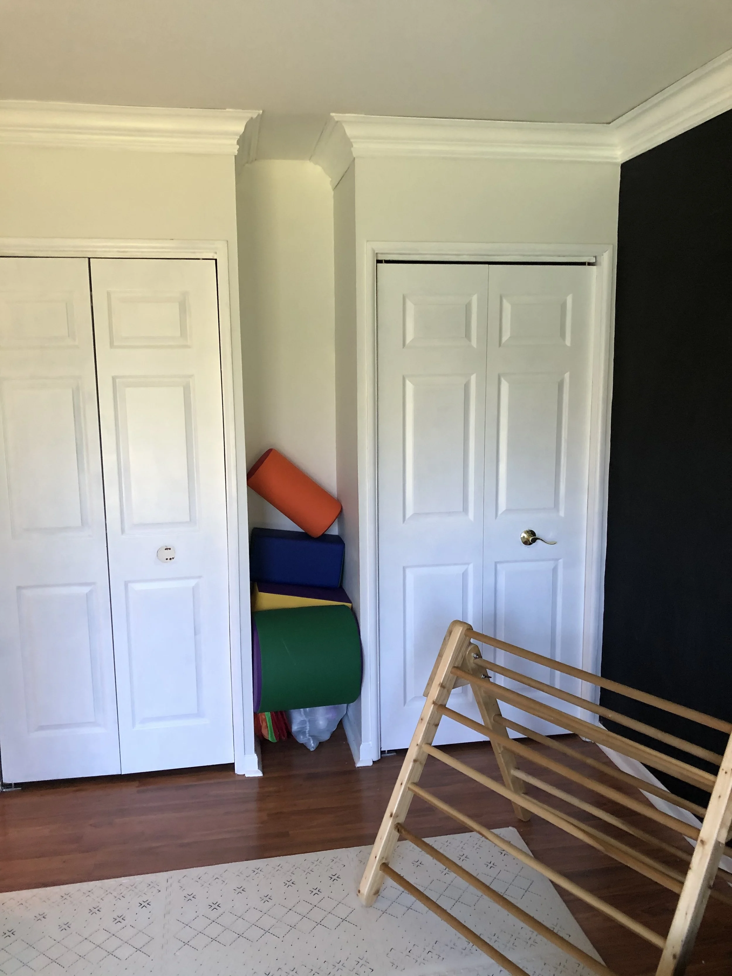 A room with white closet doors, a black accent wall, colorful foam rollers stored in a corner, a wooden drying rack, and a light-colored rug on the floor.