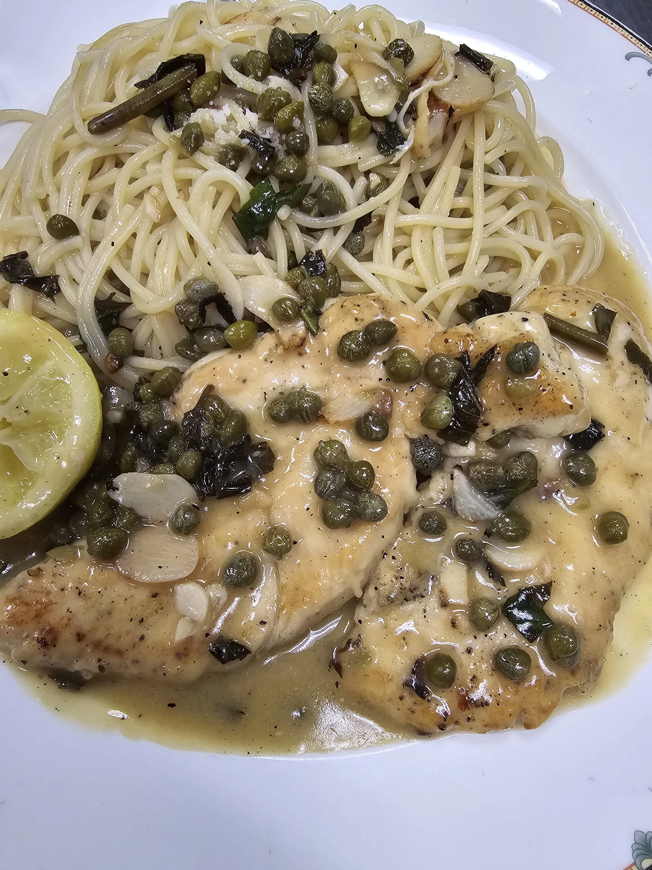 Plate of cooked chicken breast with black pepper, topped with green peppercorns, side of spaghetti pasta with green peppercorns and vegetables, and a lemon wedge.