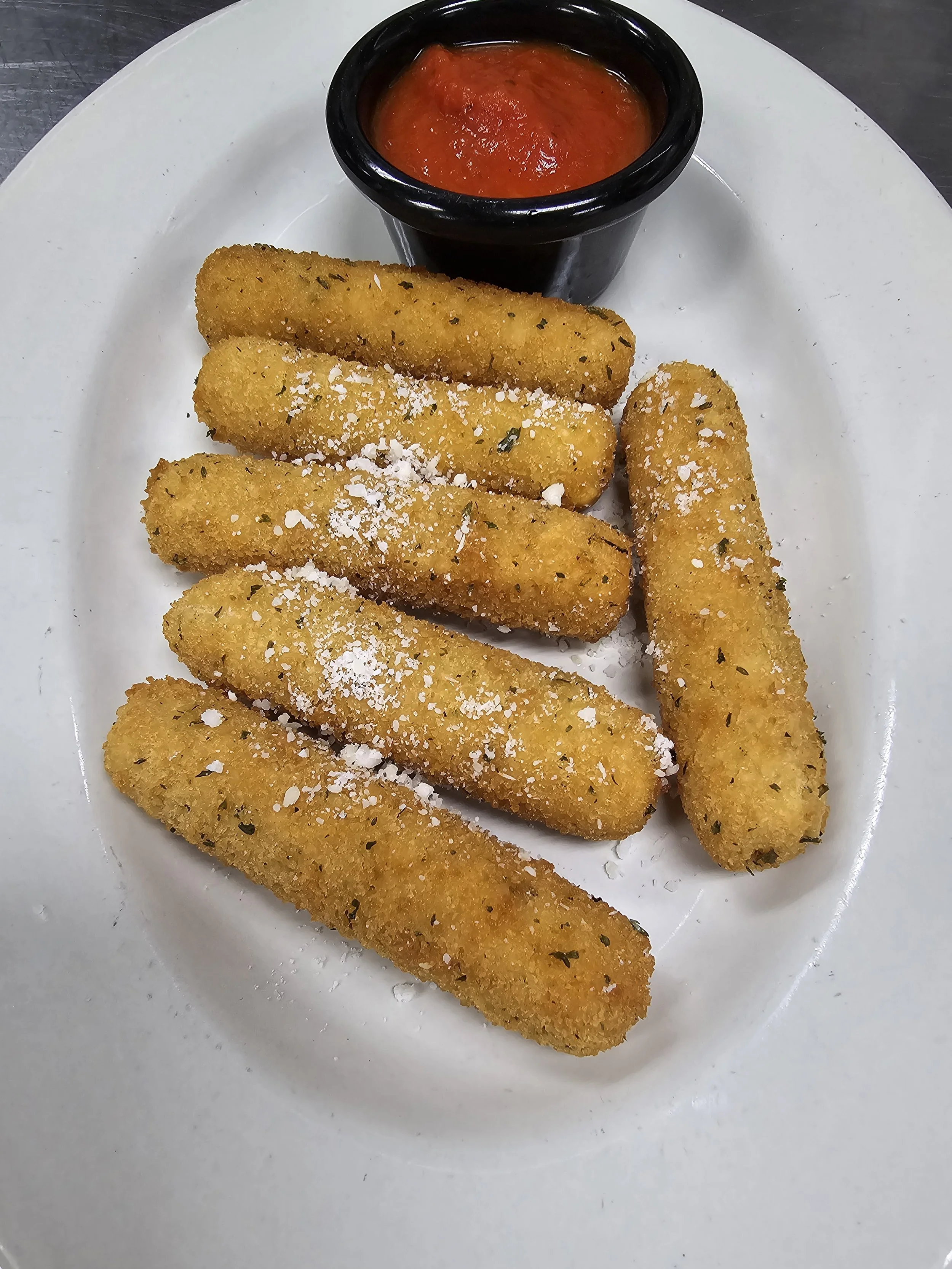 A white plate with six breaded mozzarella sticks topped with grated cheese, alongside a small black cup of marinara sauce.