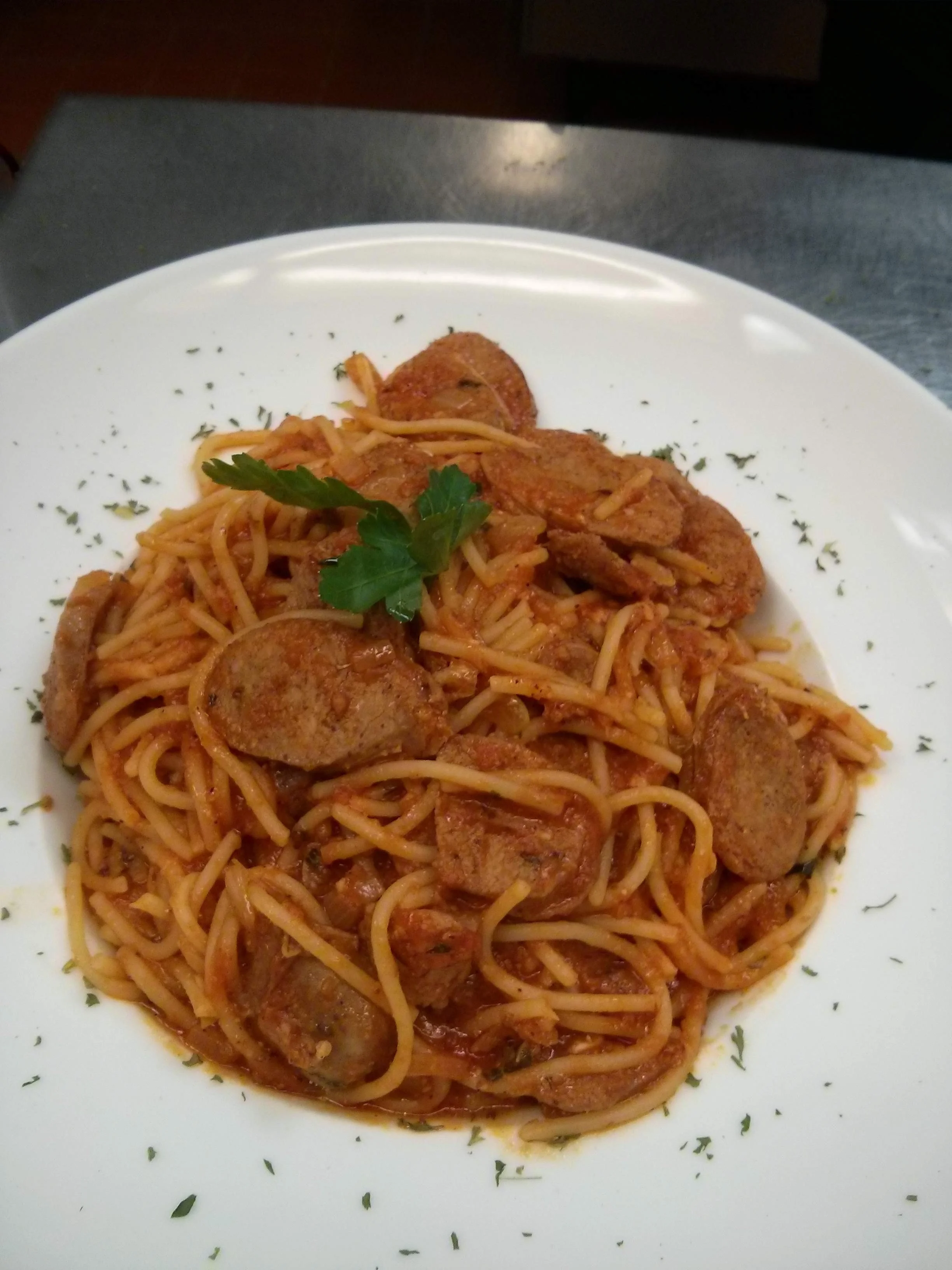 Spaghetti with sausage slices and tomato sauce garnished with parsley on a white plate.