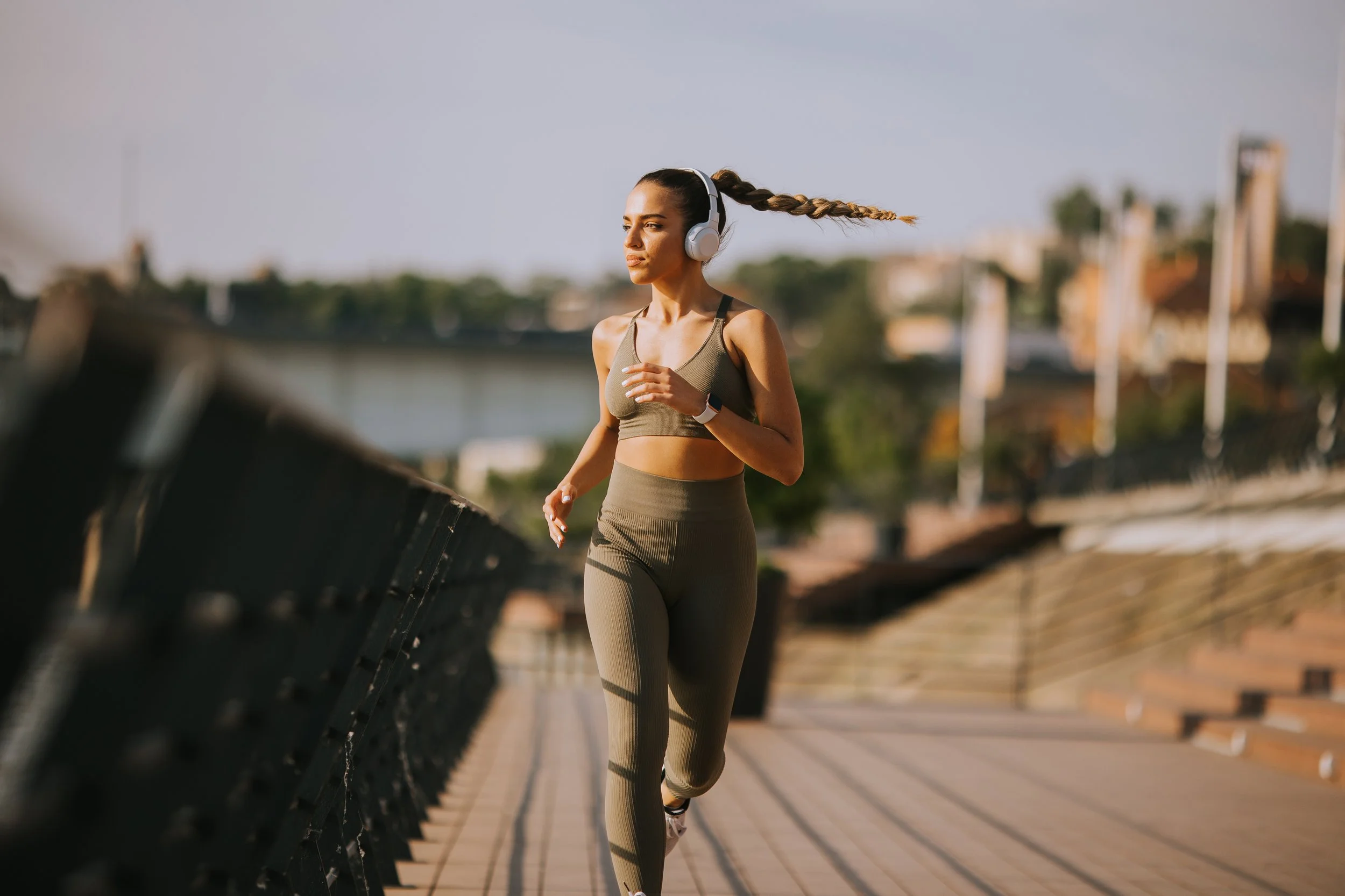 A woman jogging outdoors on a bridge, wearing workout clothing and wireless headphones, with a cityscape in the background.