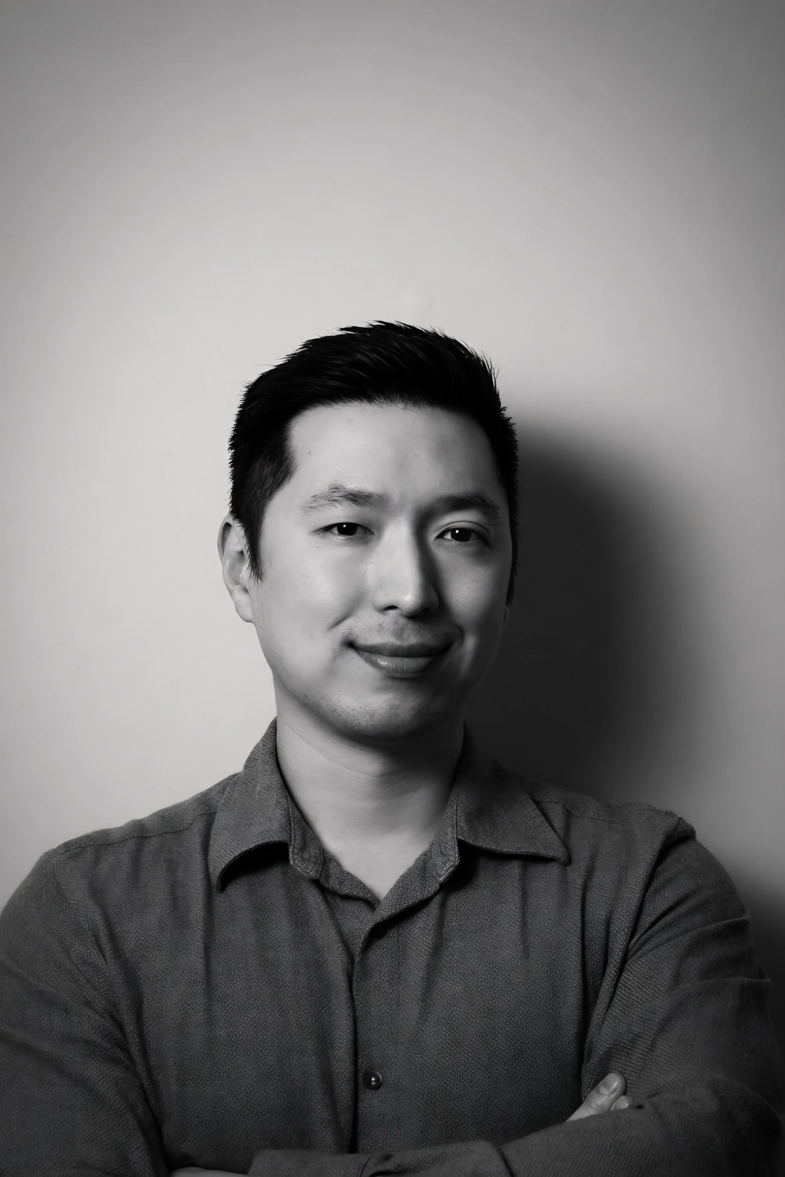A black and white portrait photograph of a young Asian man with short, slicked-back hair, wearing a collared shirt with the top button undone. He is smiling slightly and looking directly at the camera with his arms crossed in front of him.