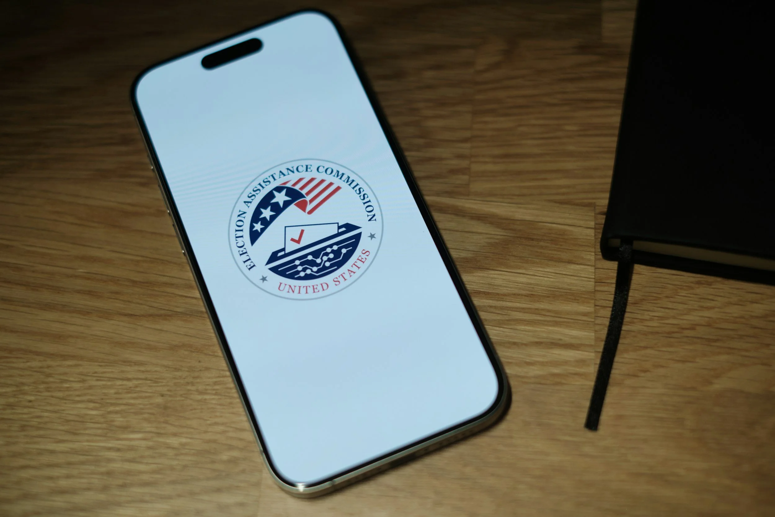 Smartphone displaying the logo of the Election Assistance Commission, positioned on a wooden surface next to a black electronic device with a cord.