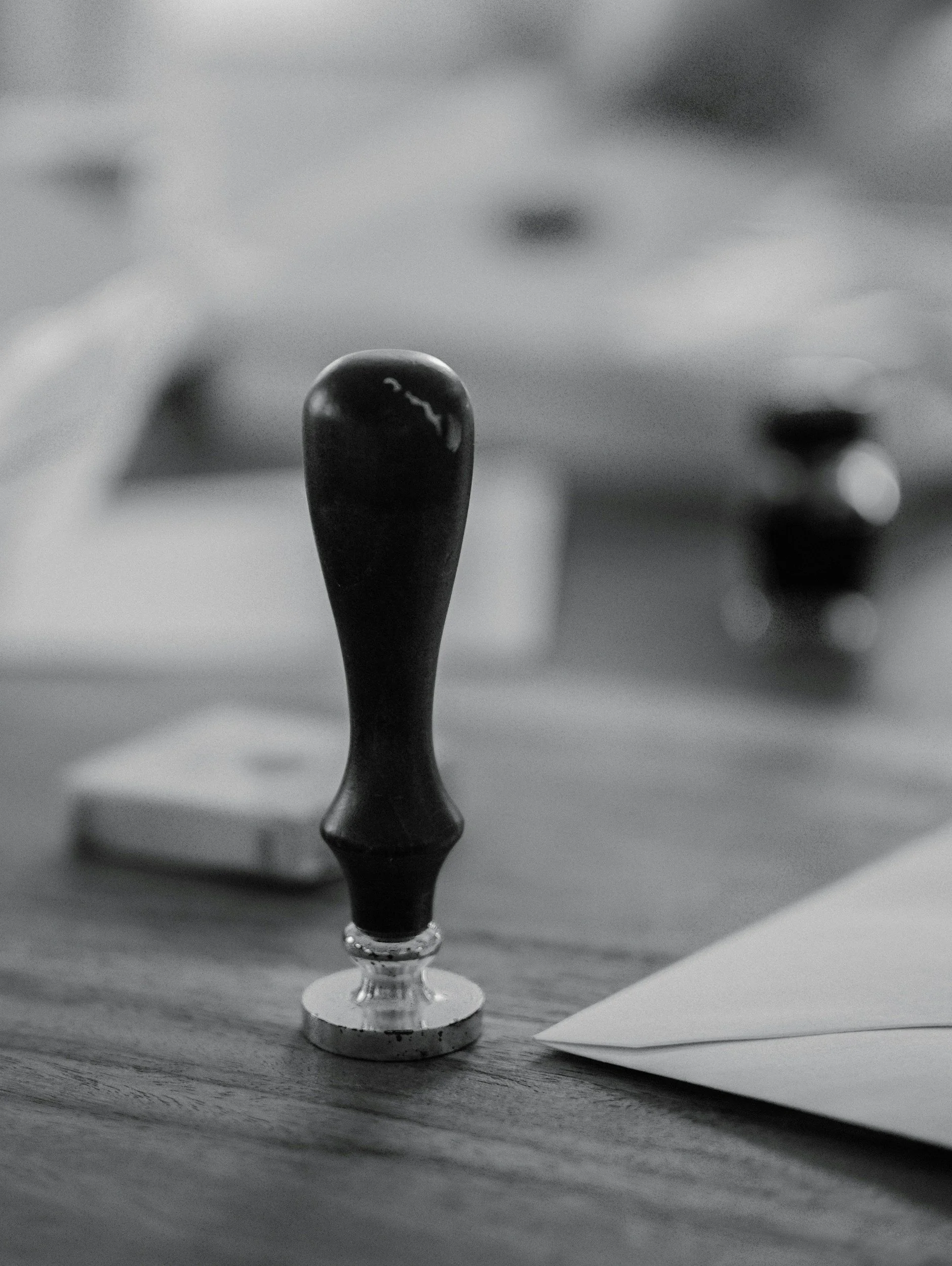 A black and white photo of a glass standing on a wooden table, with a dark substance inside and a drop of liquid at its tip. The glass is placed on a circular metallic base with paper and other objects partially visible nearby.
