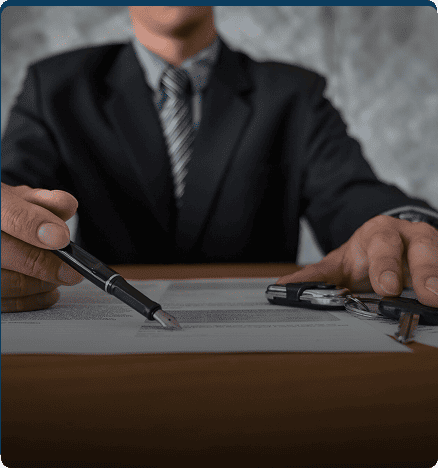 Man in suit signing a document with a pen, with keys on the table.