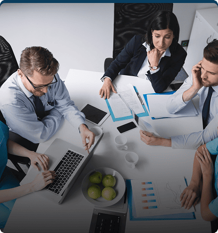 A business meeting with five people sitting around a table, discussing documents and working on laptops.