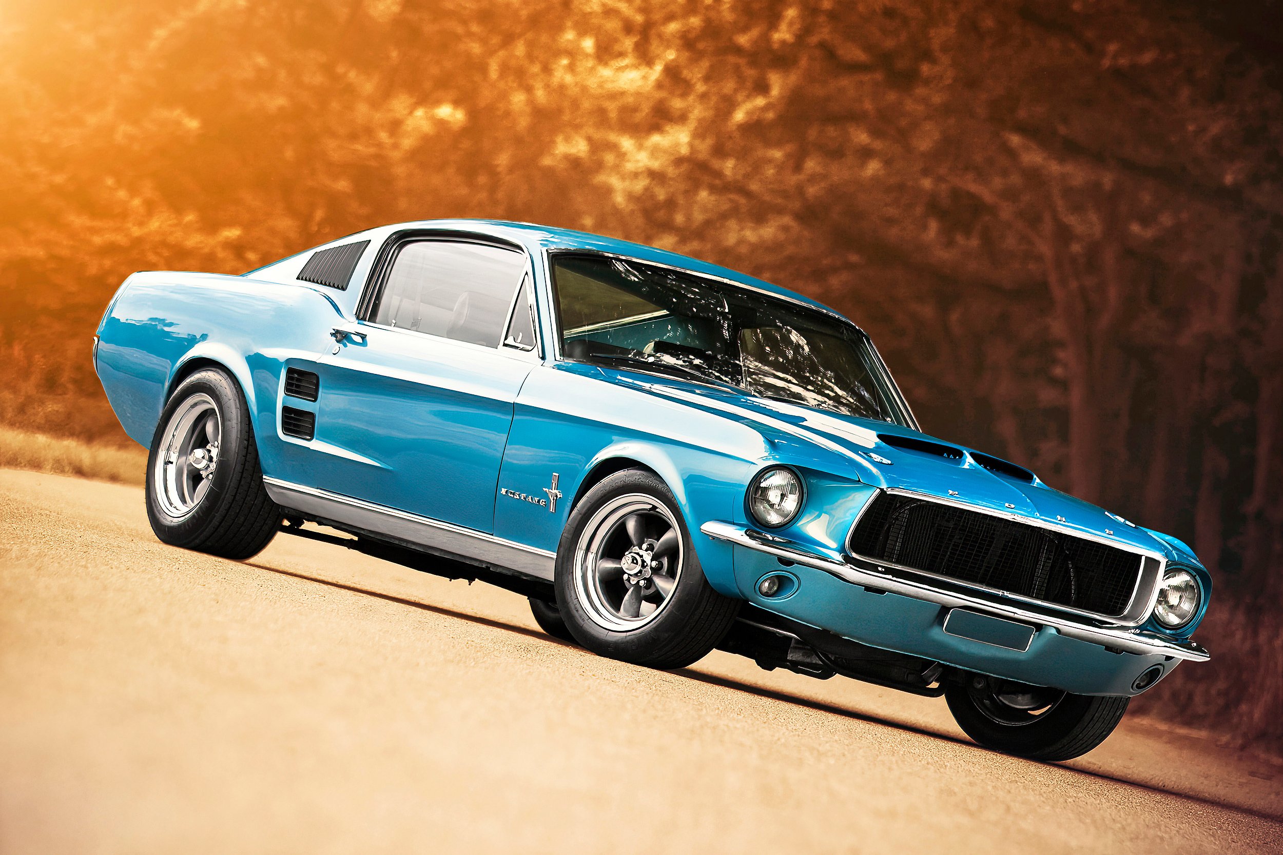 Klassischer blauer Ford Mustang Shelby GT aus den 1960er Jahren fährt auf einer Landstraße, im Hintergrund Bäume mit orangefarbenem Laub bei Sonnenuntergang.