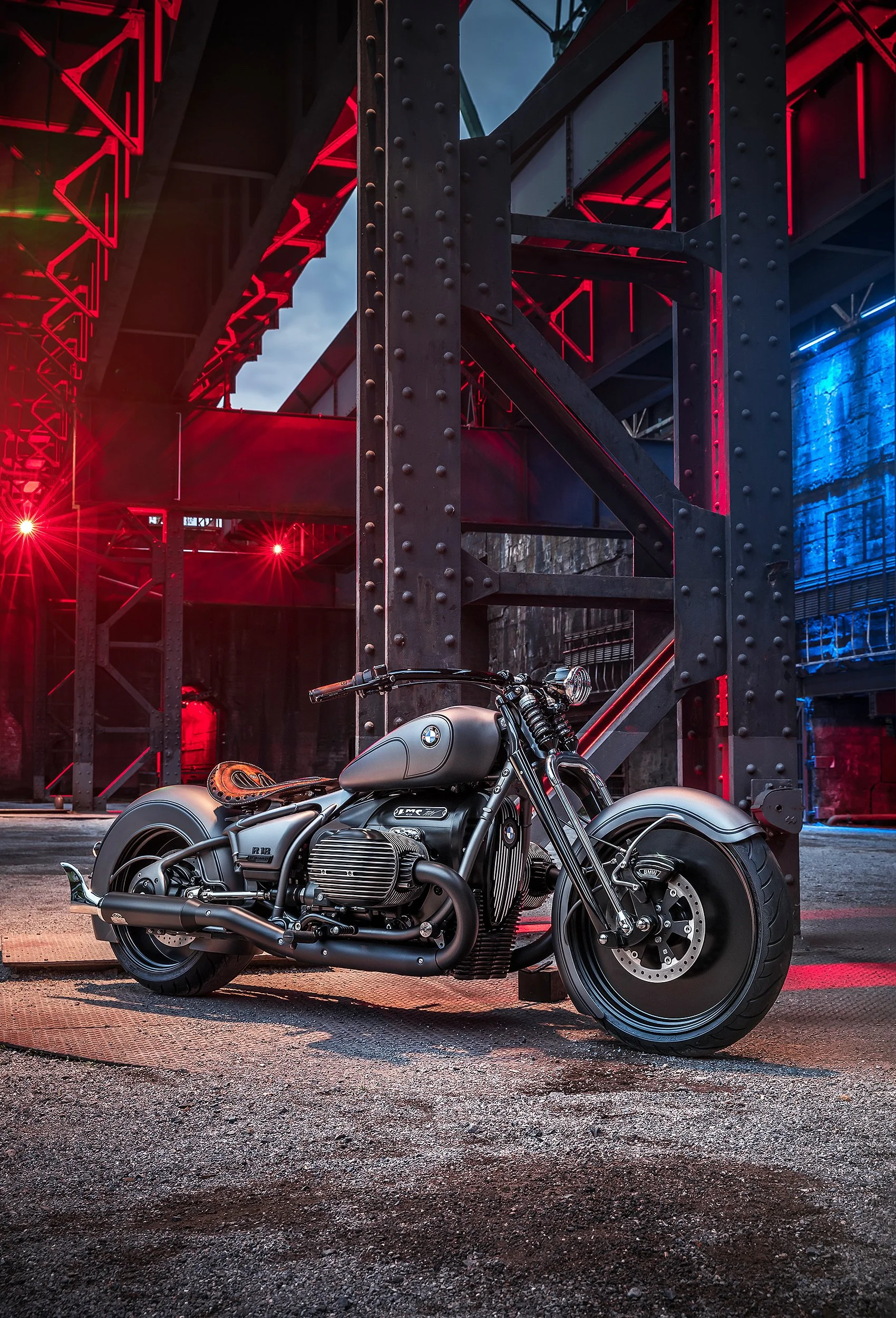 Ein schwarzes Motorrad mit BMW-Logo auf der Seite steht auf einer asphaltierte Fläche vor einer industriellen Metalltreppe mit roten und blauen Lichtern, bei Dämmerung.