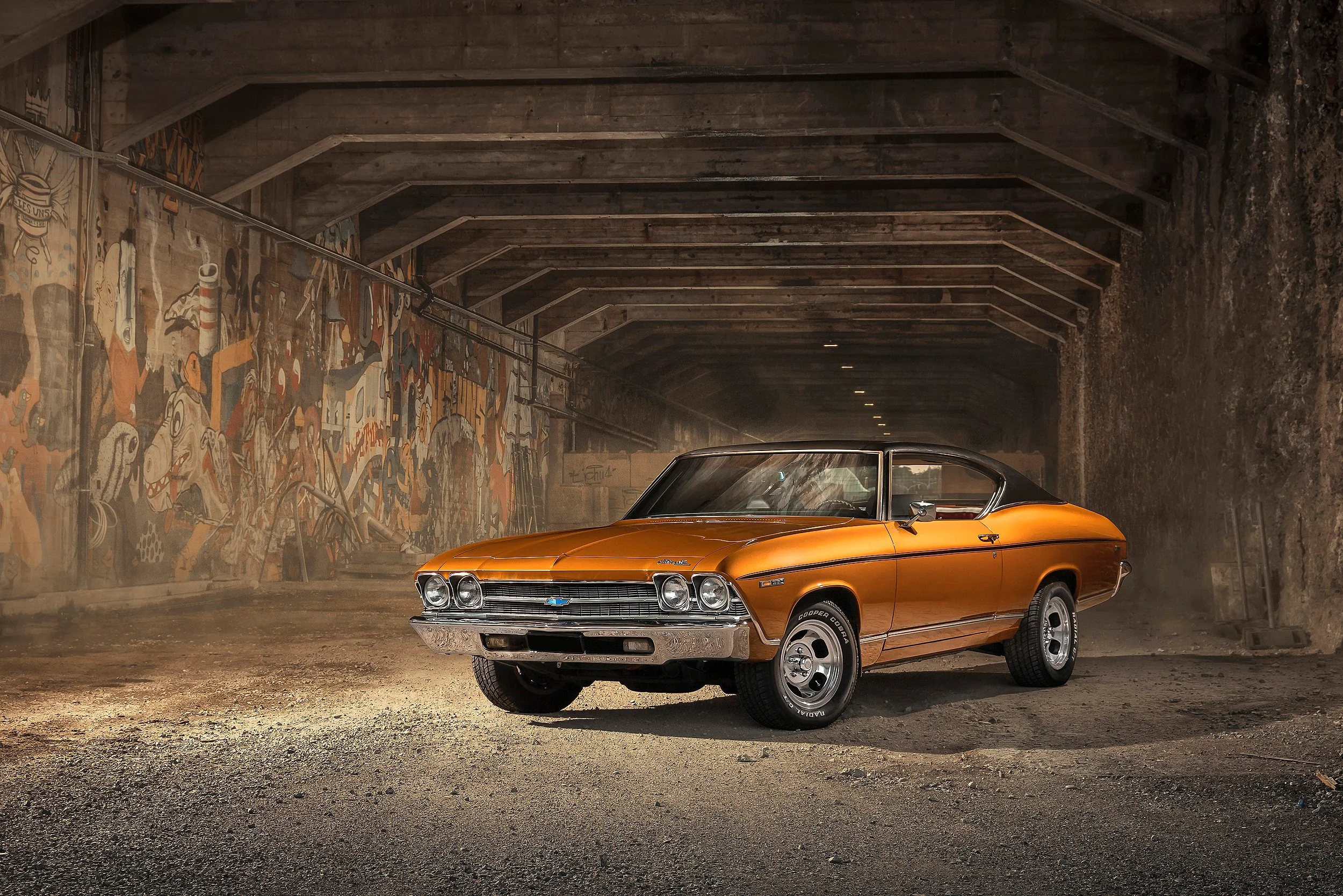 Ein orangefarbener Oldtimer-Chevrolet in einer Tunnelhalle mit Graffiti-Wand und Holzdach.