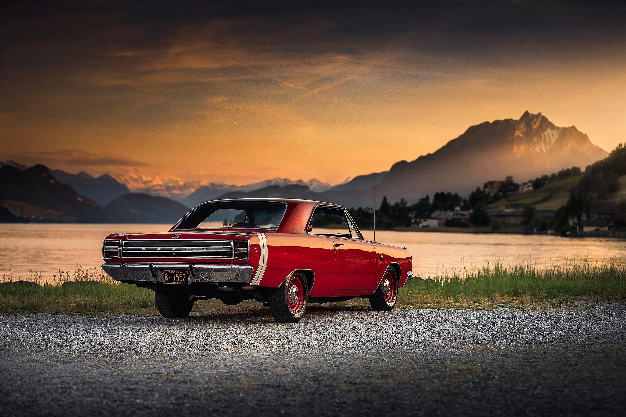 Ein roter, klassischer Oldtimer ist am Abend in einer natürlichen Umgebung mit Bergen im Hintergrund geparkt.