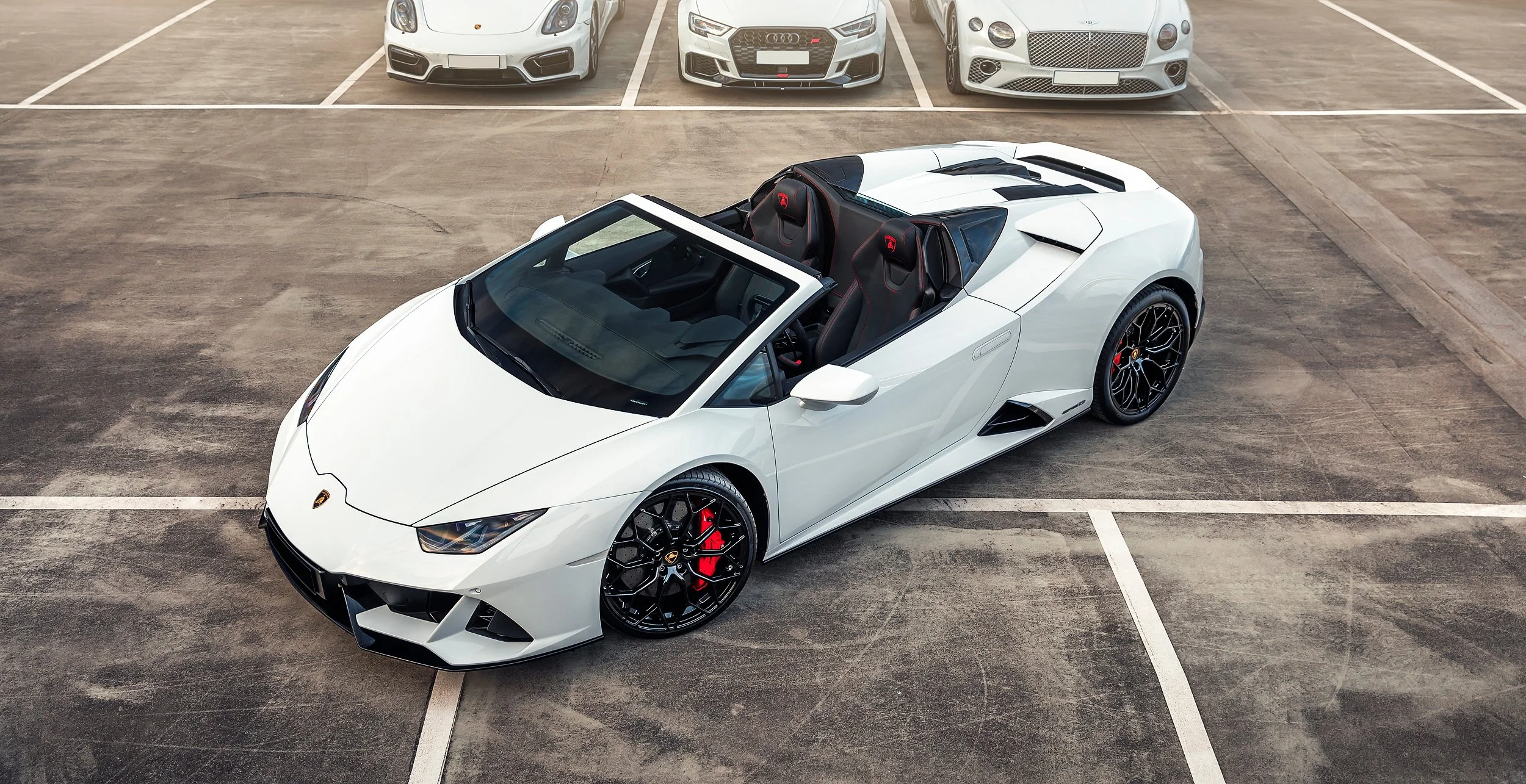 Weiße Lamborghini Cabrio mit roten Bremsen in einem leeren Parkplatz, im Hintergrund drei weitere Sportwagen, darunter ein Audi und ein Maserati.