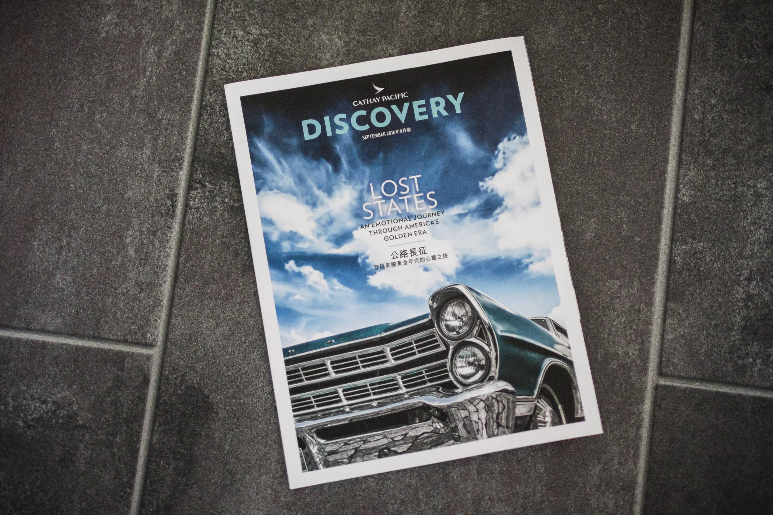 Flyer der Fluggesellschaft Cathay Pacific mit dem Titel 'Discovery' und einem Auto vor einem blauen Himmel mit Wolken.