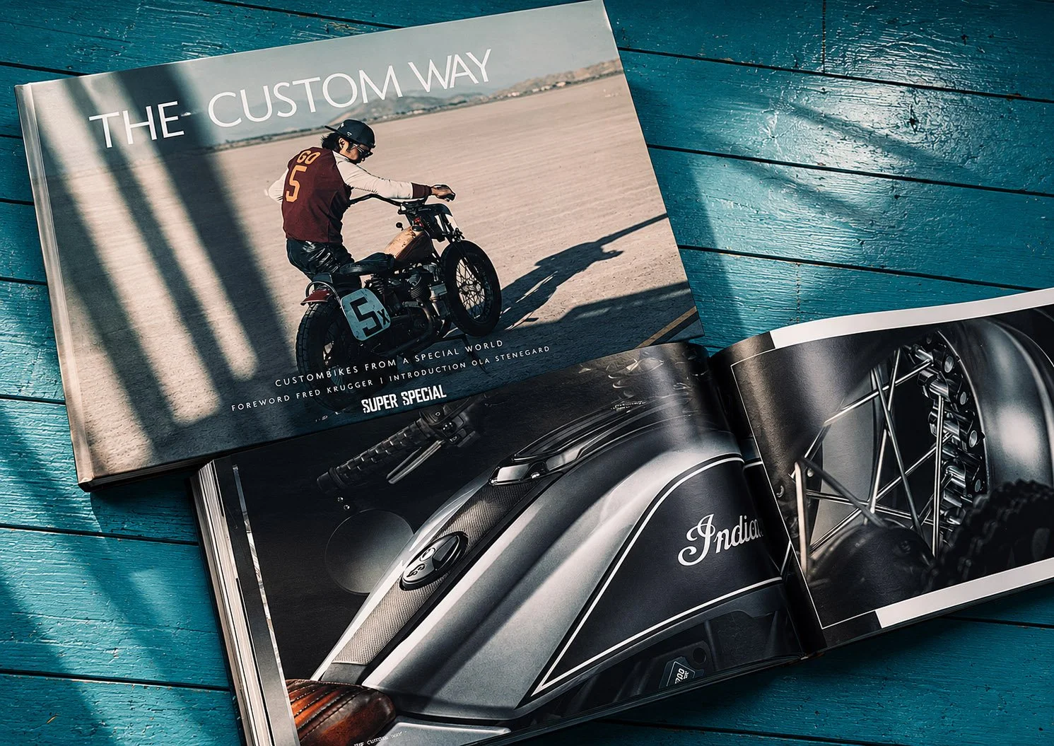 Offener Magazin auf einem blauen Holztablett, zeigt eine Seite mit einem Motorrad und dem Titel "The Custom Way", die andere Seite zeigt ein Detail eines Motorrads mit der Marke "Indian".