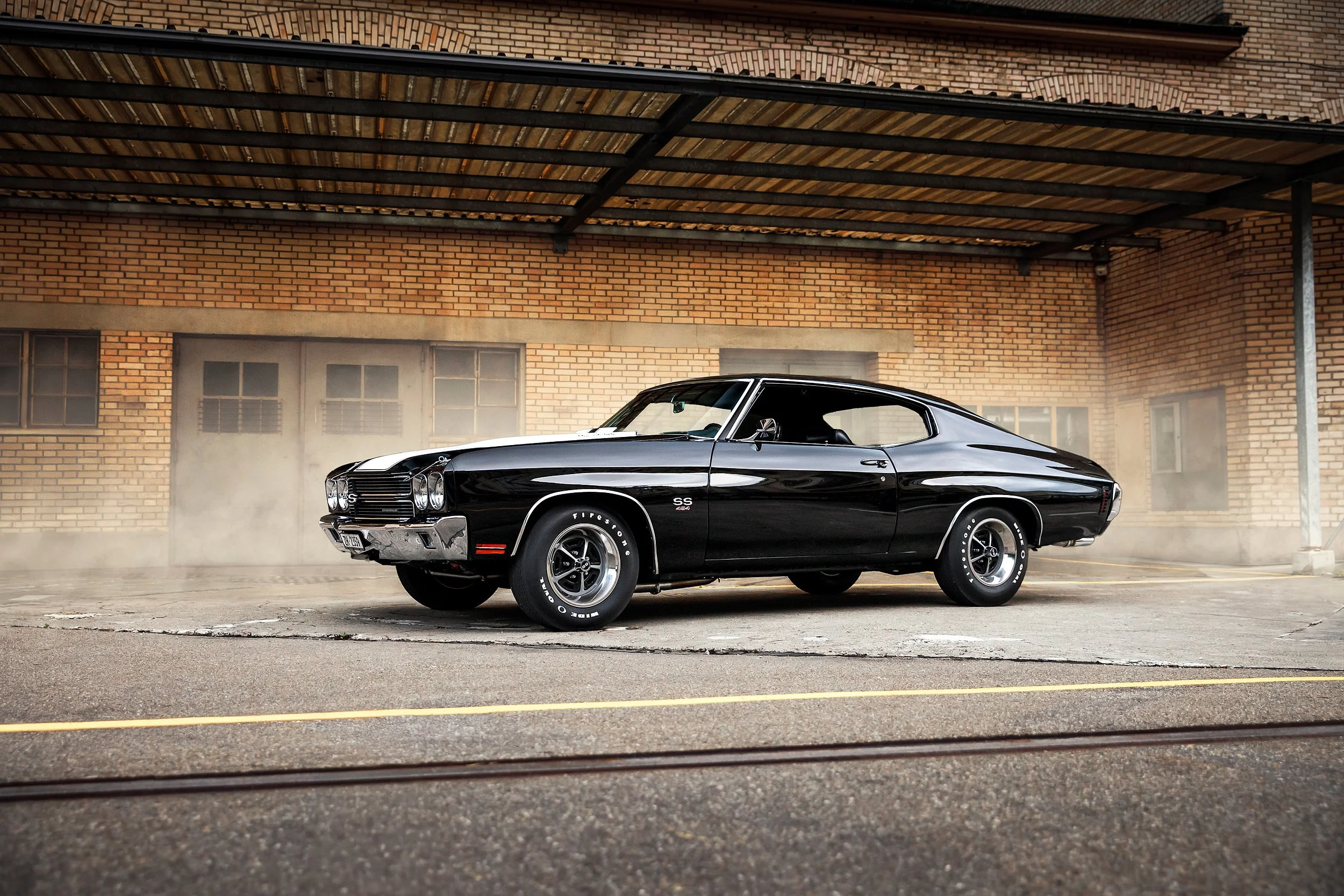 Ein schwarzer Oldtimer-Chevrolet Camaro SS 454 auf einem Parkplatz vor einer Backsteinwand, sichtbar durch eine Scheibe mit Rahmen.