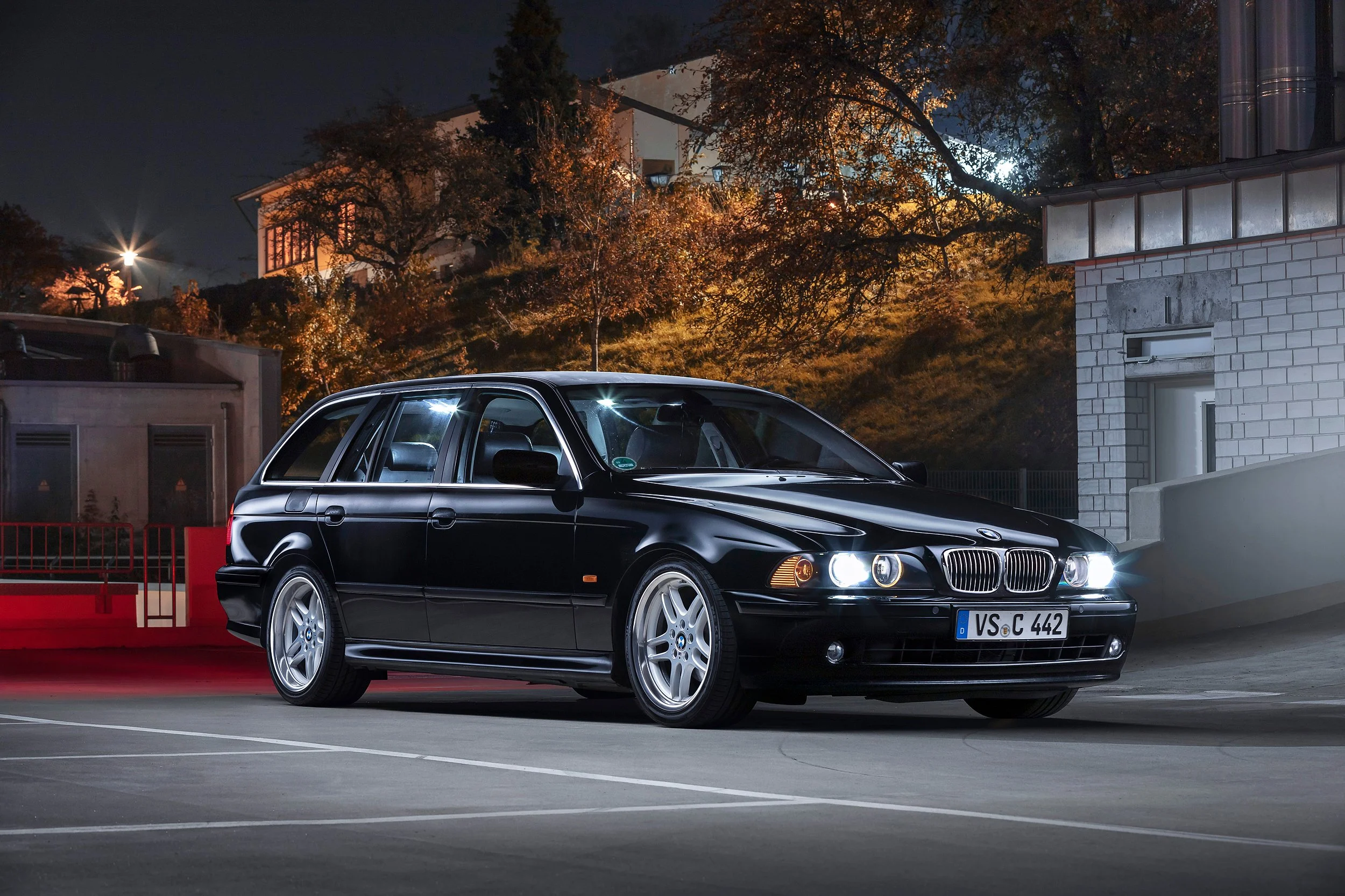 Schwarzer BMW E34 5er Modell mit deutschem Nummernschild, parkt in einer leeren Parkgarage bei Nacht, im Hintergrund sind Bäume und Gebäude mit Beleuchtung.