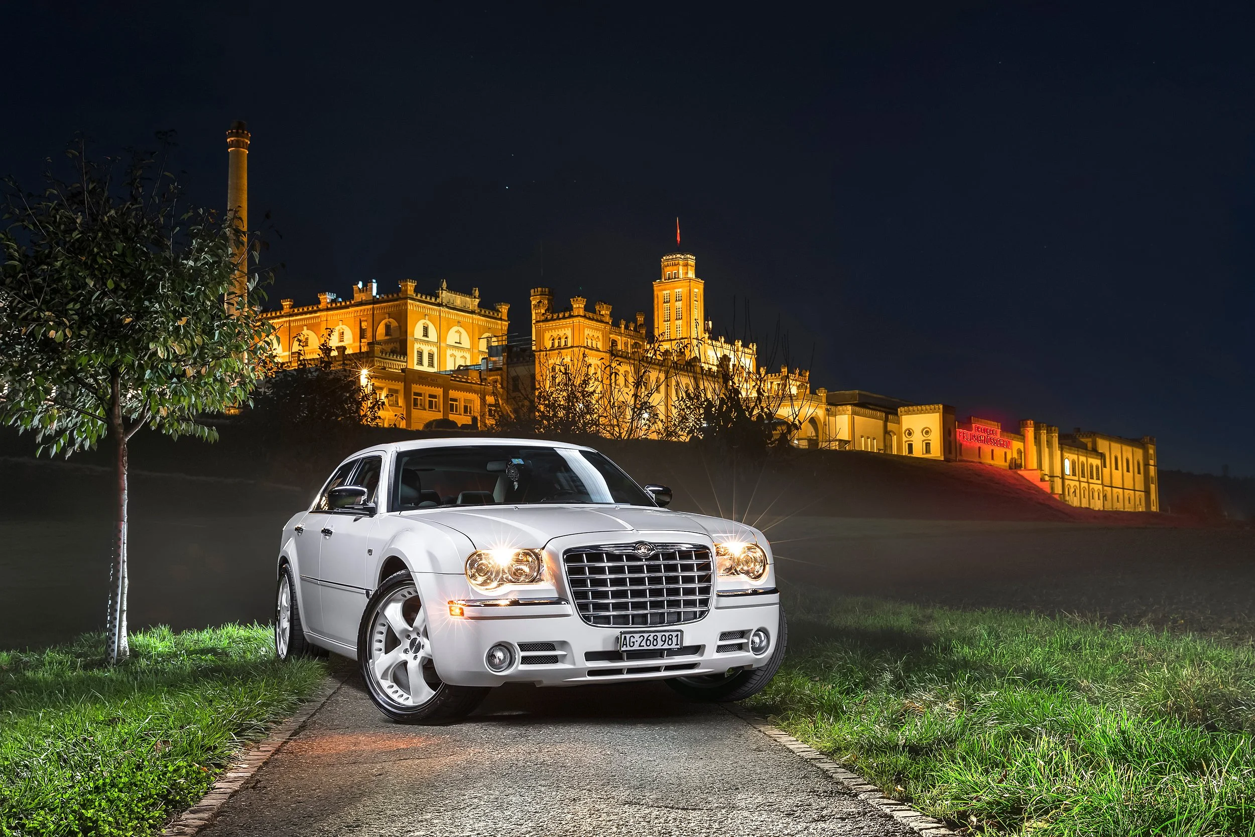 Ein weißer Chrysler 300C steht auf einer ruhigen Straße vor einer grasbewachsenen Fläche, im Hintergrund ein majestätisches, beleuchtetes Schloss bei Nacht, links ein Baum, der den Himmel überragt.
