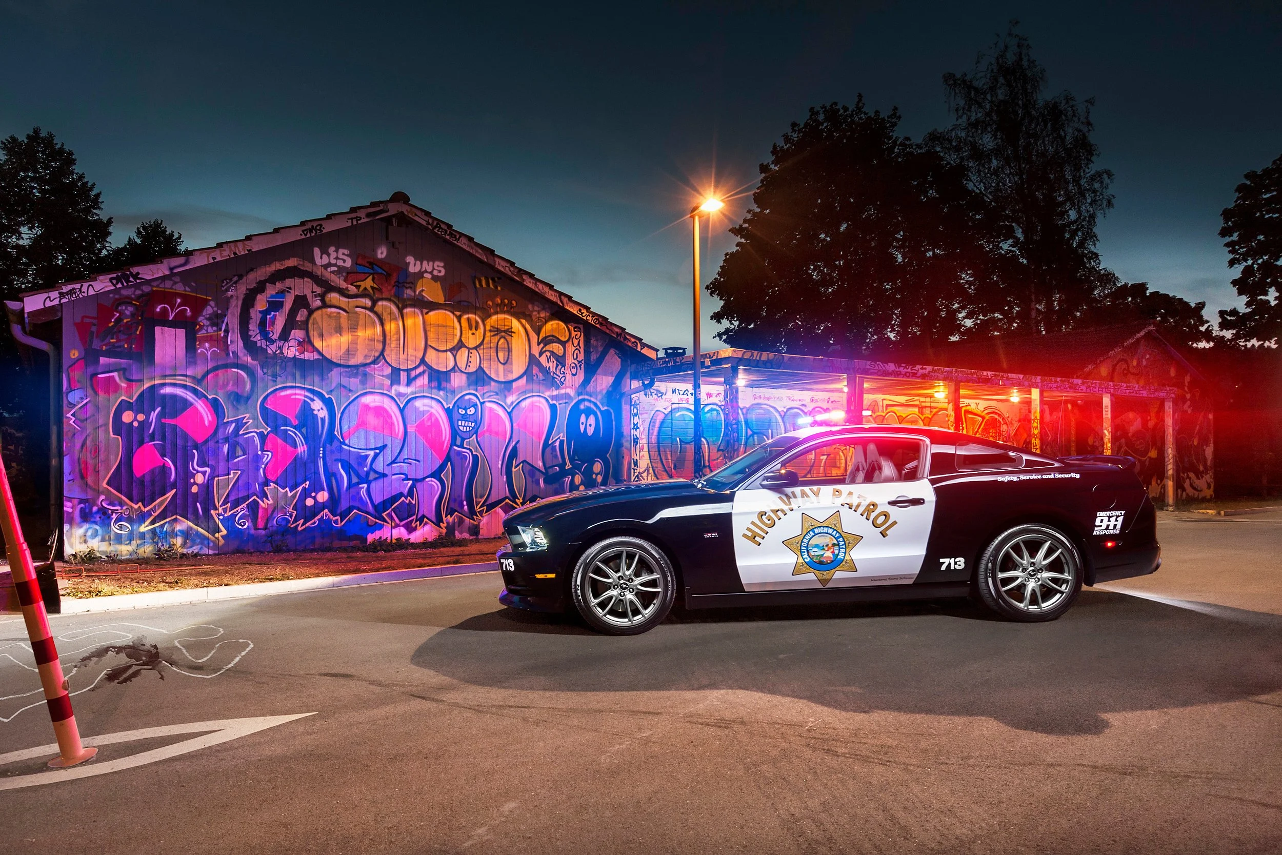 Ein Polizeiwagen der Highway Patrol steht in einem beleuchteten Parkplatz bei Nacht, hinter einem mit Graffiti bemalten Gebäude und Bäumen.