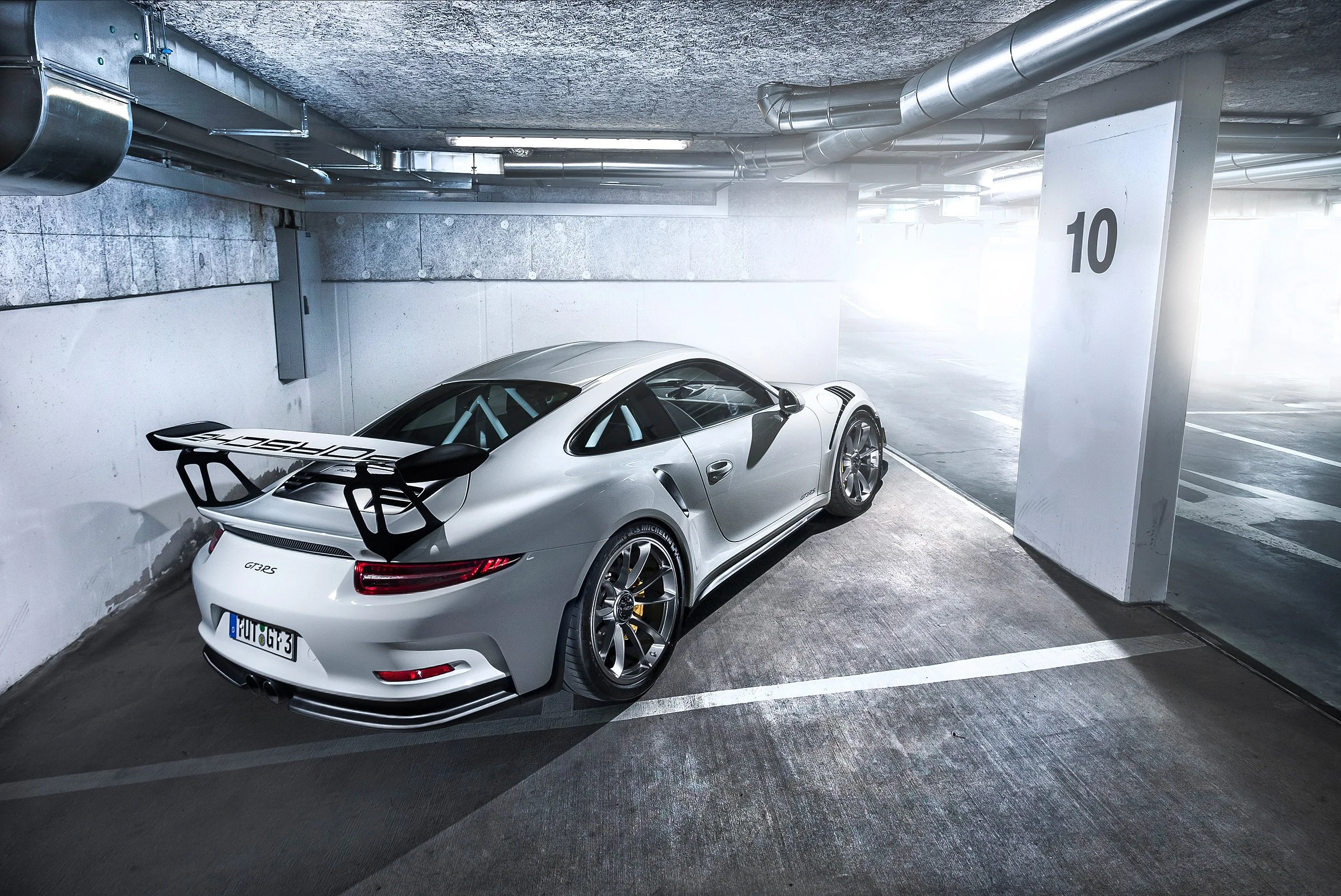Weißer Porsche 911 GT3 RS mit großem Heckspoiler steht in einer Parkgarage neben einer weißen Säule mit der Nummer 10, umgeben von grauen Wänden und Decken, mit sichtbaren Rohren und Kabeln.