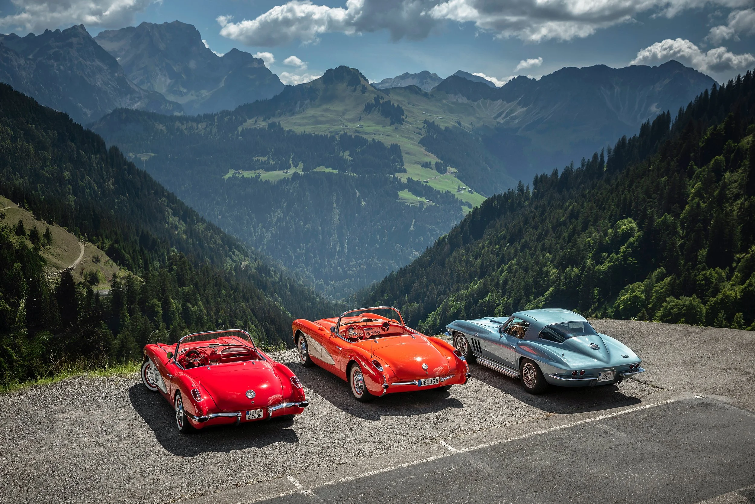 Fünf klassische Sportwagen, darunter drei rote und zwei silberne, parken in einer Bergregion mit grünen Hängen und hohen Bergen im Hintergrund, bei bewölktem Himmel.