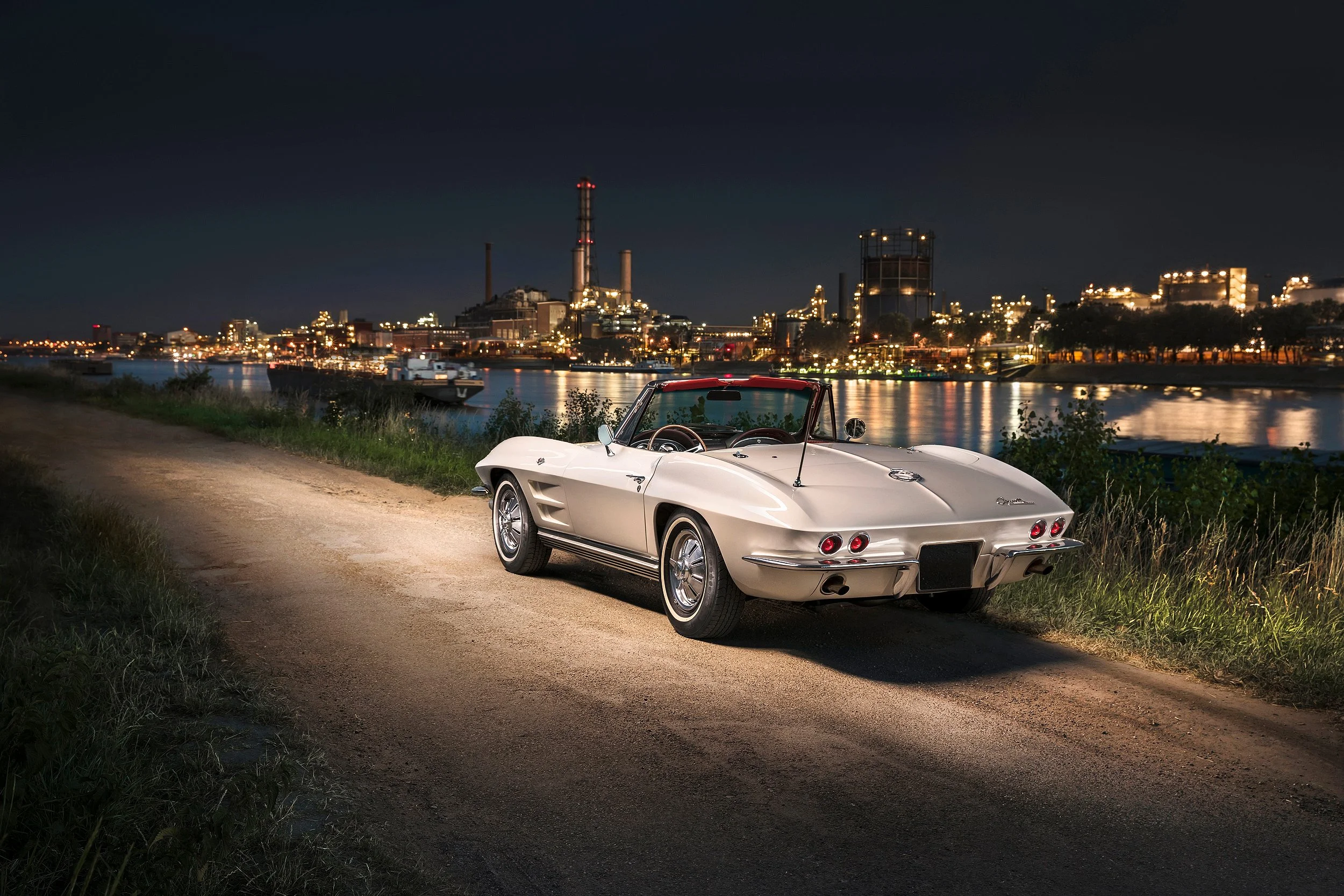 Ein weißer Oldtimersportwagen parkt am Ufer bei Nacht, mit einer industriellen Stadt im Hintergrund, die an beleuchteten Gebäuden und einer Fabrikanlage erkennbar ist.