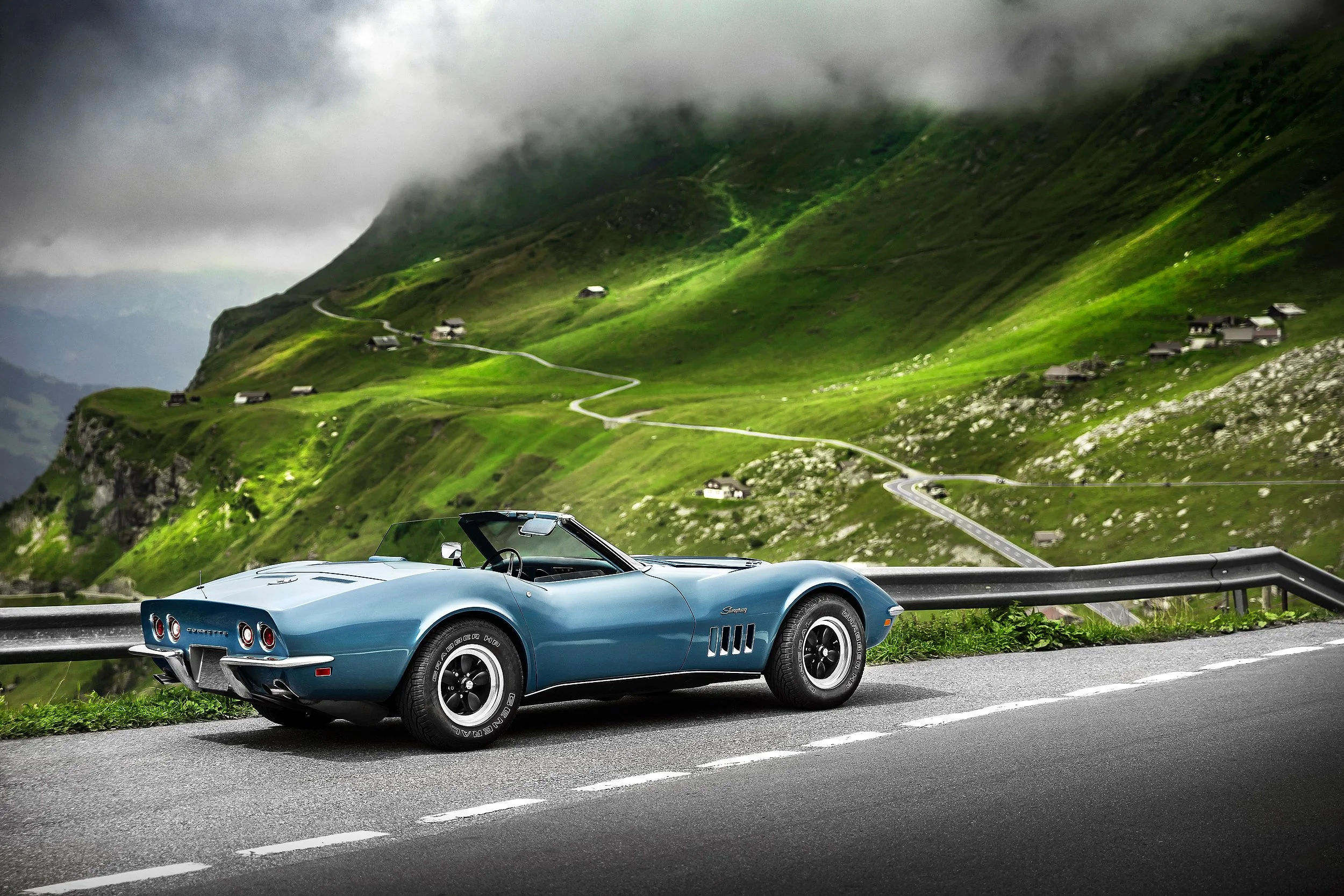 Ein blauer Oldtimer-Cabrio fährt auf einer Serpentine durch eine grüne Berglandschaft. Im Hintergrund hängen Wolken über den Hügeln.